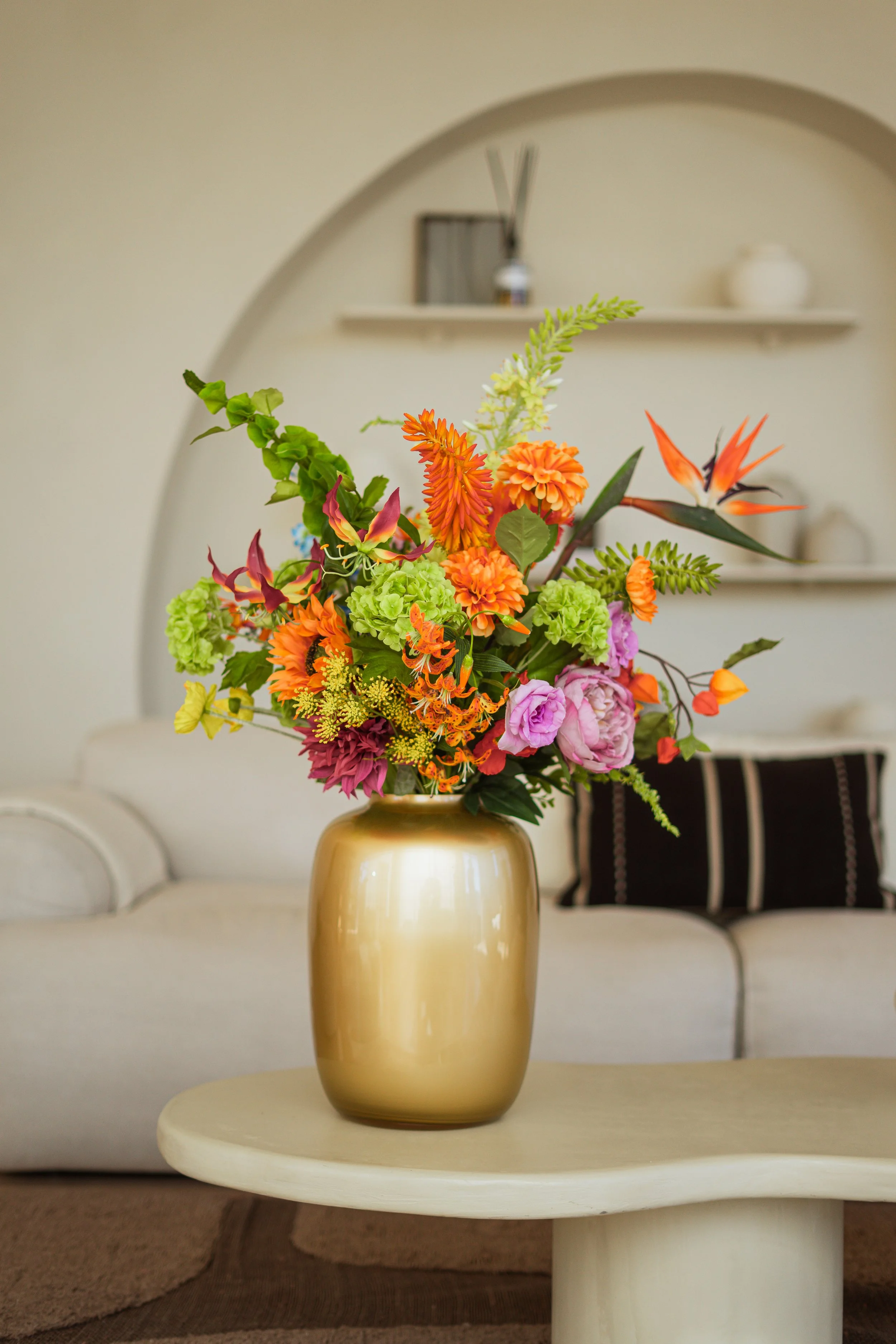 Een bloemenvaas met kleurrijke bloemen op een ronde, lichte tafel in een woonkamer met een bank en decoratieve planken op de achtergrond. Floweris