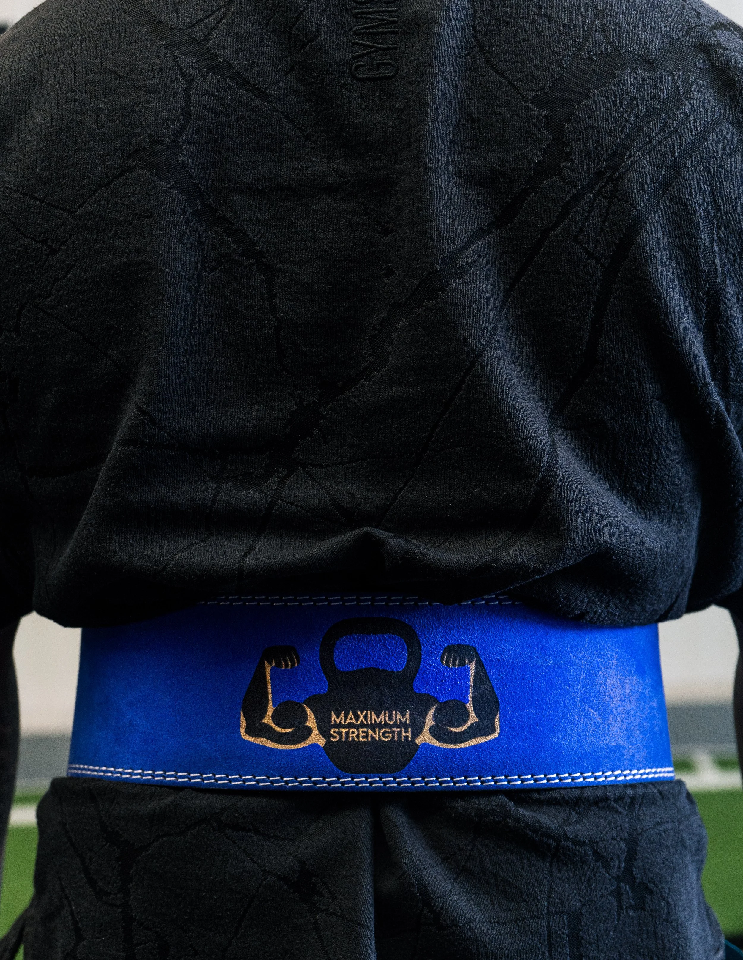 Close-up van een persoon in een zwart sportoutfit met een blauwe riem met de tekst 'Maximum Strength' en een buideldbare musculaire arm in zwart en geel