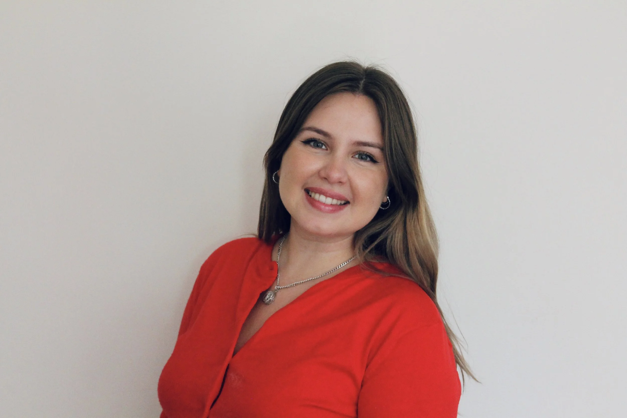 Image of a smiling caucasian woman, with green eyes and long, straight light brown hair. She is wearing winged eyeliner and lip gloss, and a red cardigan with a silver locket and silver earrings.
