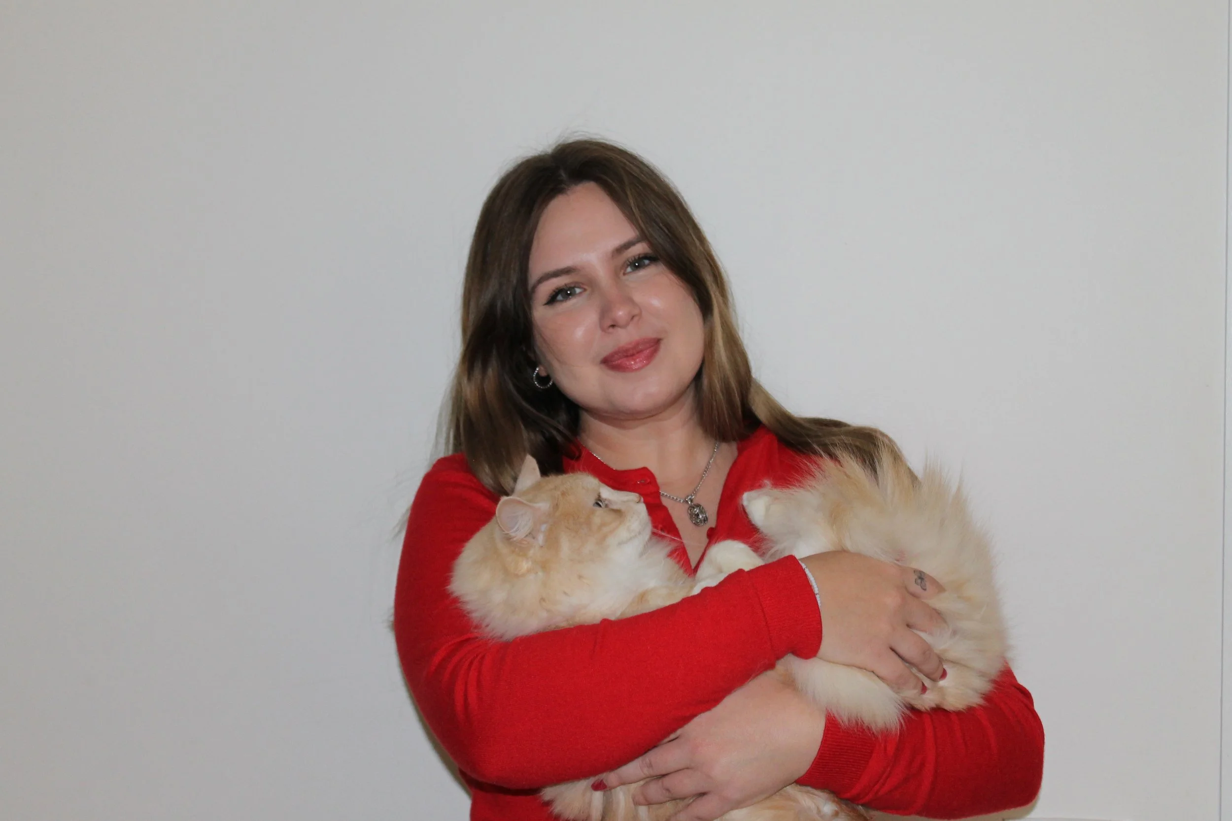 Annabelle smiles at the camera while holding her blond longhair cat. She is wearing a red top against a plain background.
