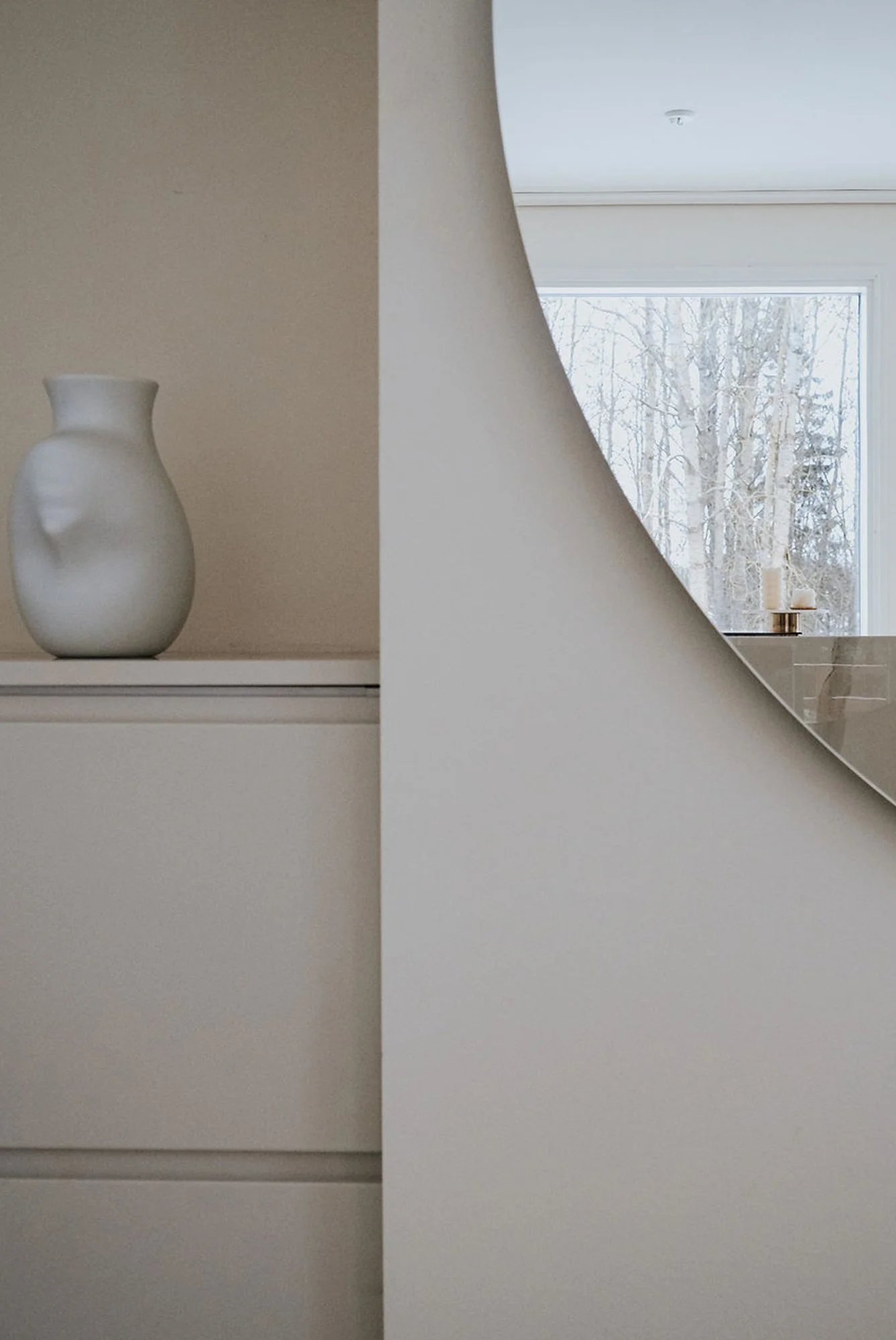 Eteistila vaalealla tasolla, jonka päällä on valkoinen maljakko, minimalistinen sisustus. Osittain peilissä heijastuneen ikkunan ja talvimaiseman näkyvissä.
