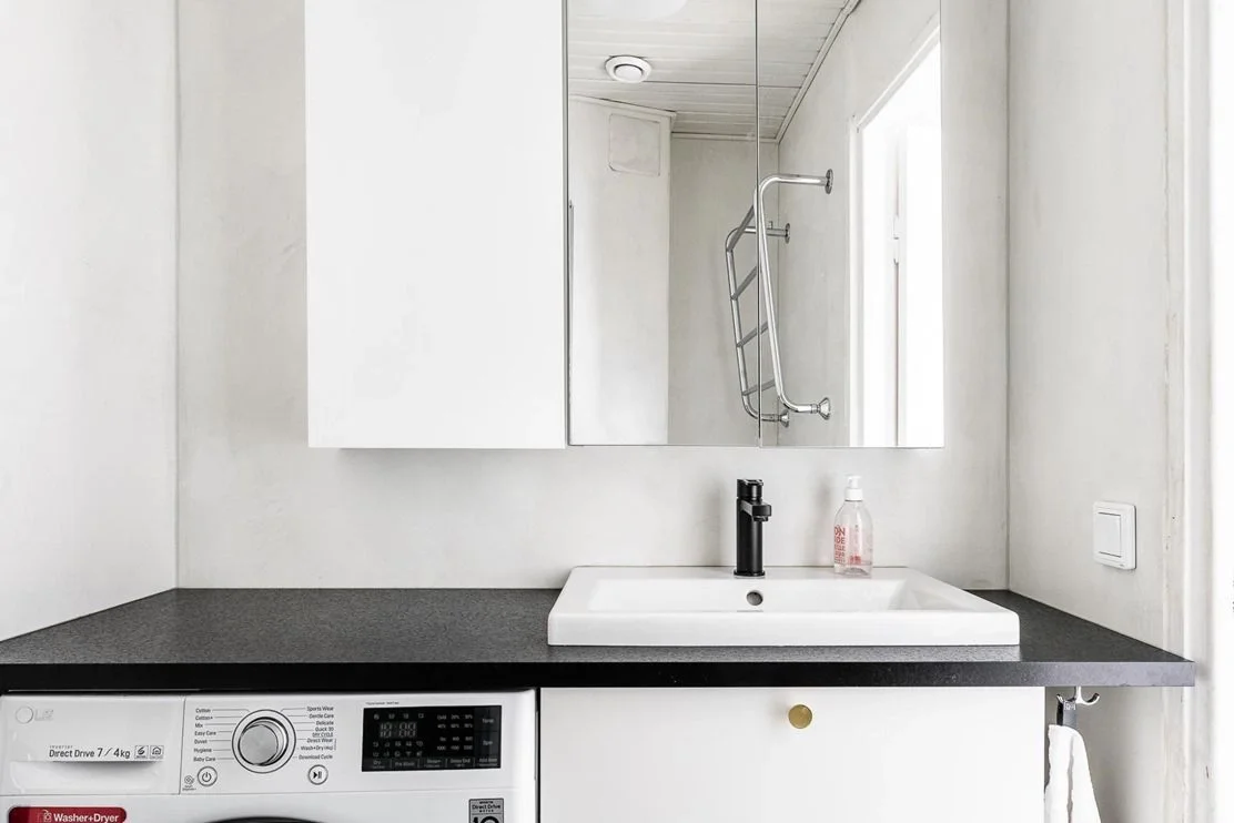 Yksinkertainen kylpyhuone, jossa on pesuallas, suuri peili ja pesukone alhaalla, pyyhkeenpidike oikealla ja käsidesipullo pöydällä.