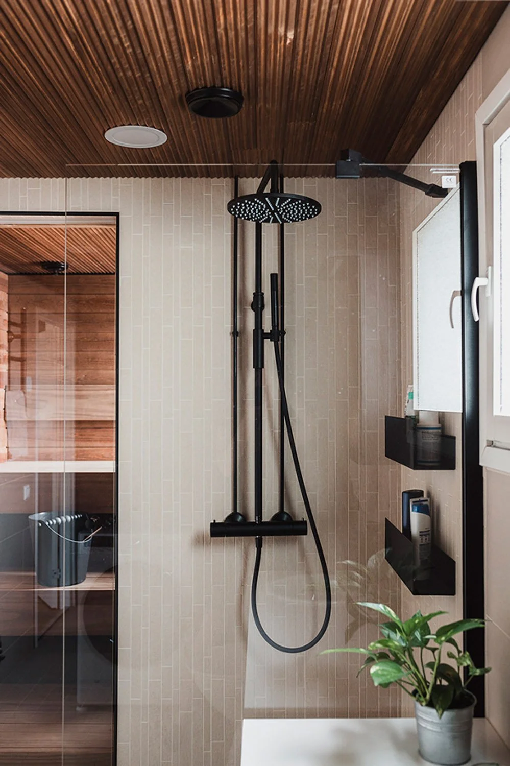 Musta suihkuhuoneen suihkukaappi ja suihkuosa, jossa musta sadesuihku ja käsisuihku, viherkasvi valkoisessa ruukussa pöydällä, keraamiset seinälaatat ja savupiippu katossa.