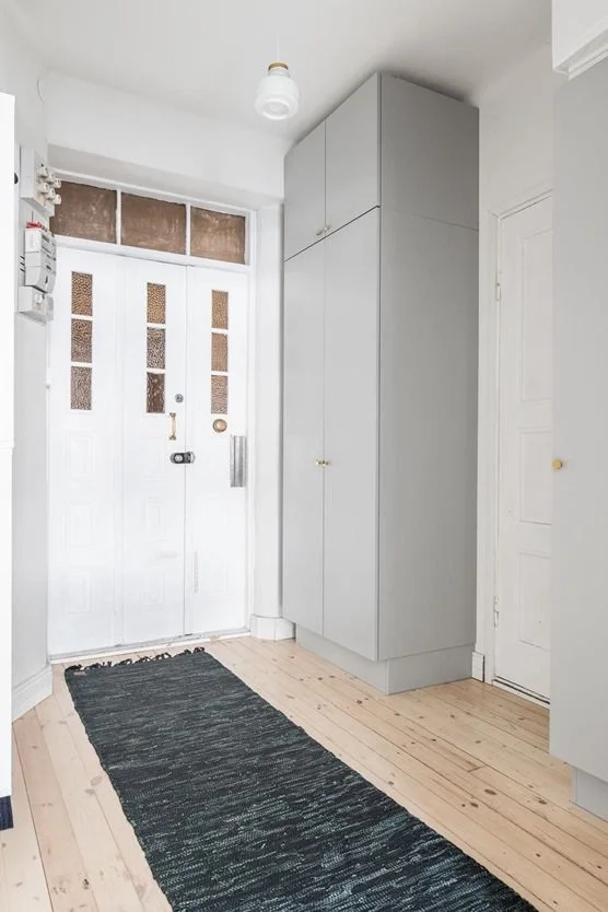 Kuvassa on eteinen, jossa on valkoinen ovi, musta matto ja suuret vaaleanharmaat kaapit