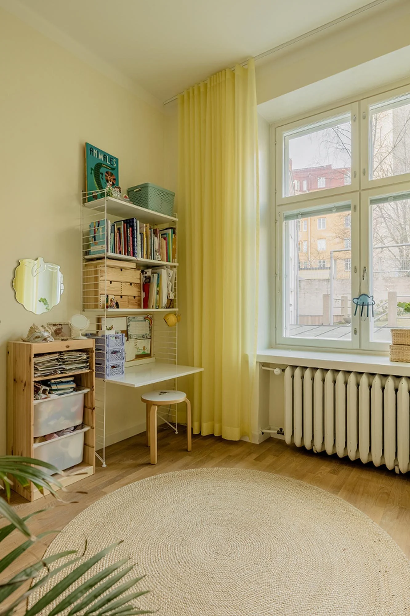 Valoisa lapsen huone, jossa keltainen verho ja matto, kirjoituspöytä ja kirjahylly, iso ikkuna, jossa ulkona rakennuksia ja puita.