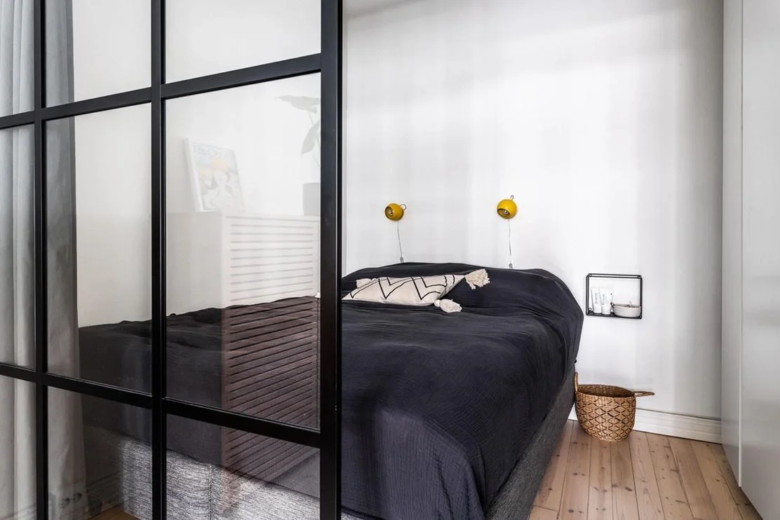 Makuuhuone, jossa musta sänky, kaksi keltaista lukulamppua seinällä, pieni seinähylly ja koria lattialla.