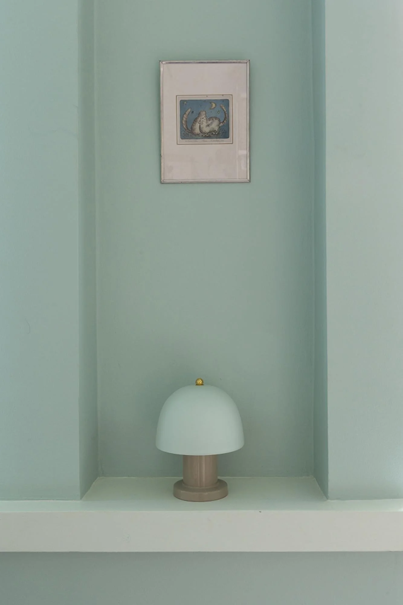 Valaiseinällä on taulukko ja pieni pöytälamppu, taulukossa on kuvia ja kukkia, huoneen seinä on vaaleanvihreä.