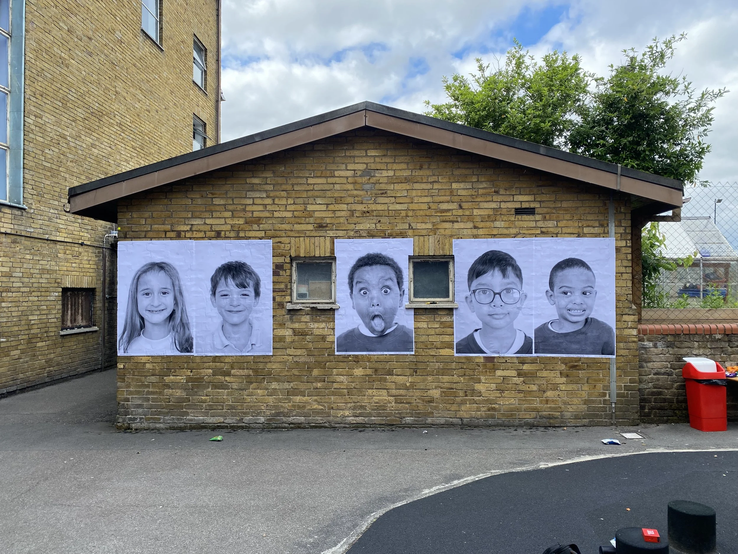 A series of large black-and-white portraits of diverse children displayed on a brick wall in an urban outdoor setting.
