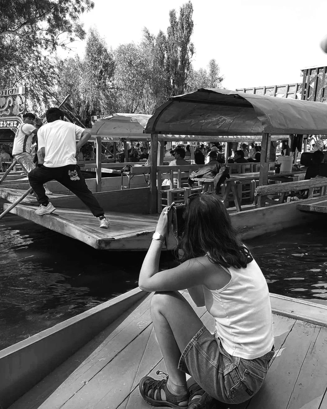 A woman sitting on a boat at a waterfront, taking a photo of another boat passing by with people on it, in a busy outdoor setting with trees and other boats.