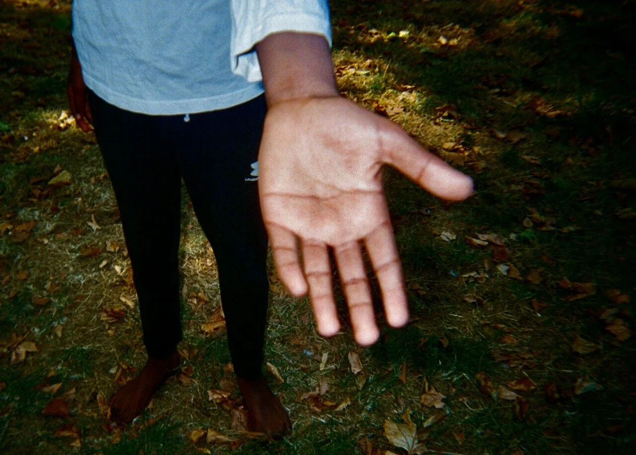Person standing barefoot outdoors, reaching out with their right hand towards the camera, background of grass and fallen leaves.