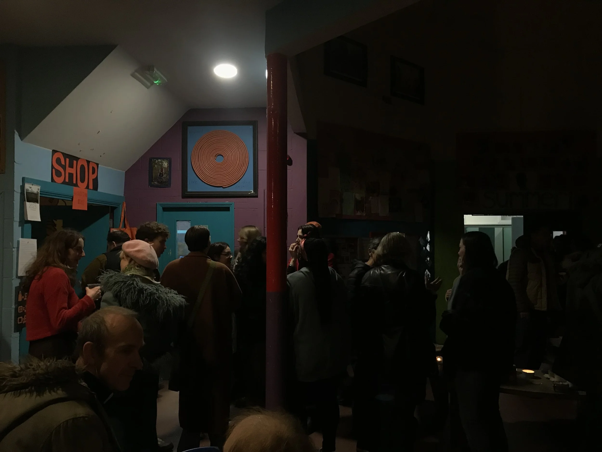 People gathered in a dimly lit indoor space, with some engaging in conversation, a brightly lit ceiling light, framed artwork on purple walls, and a sign that reads 'SHOP' on the left side.