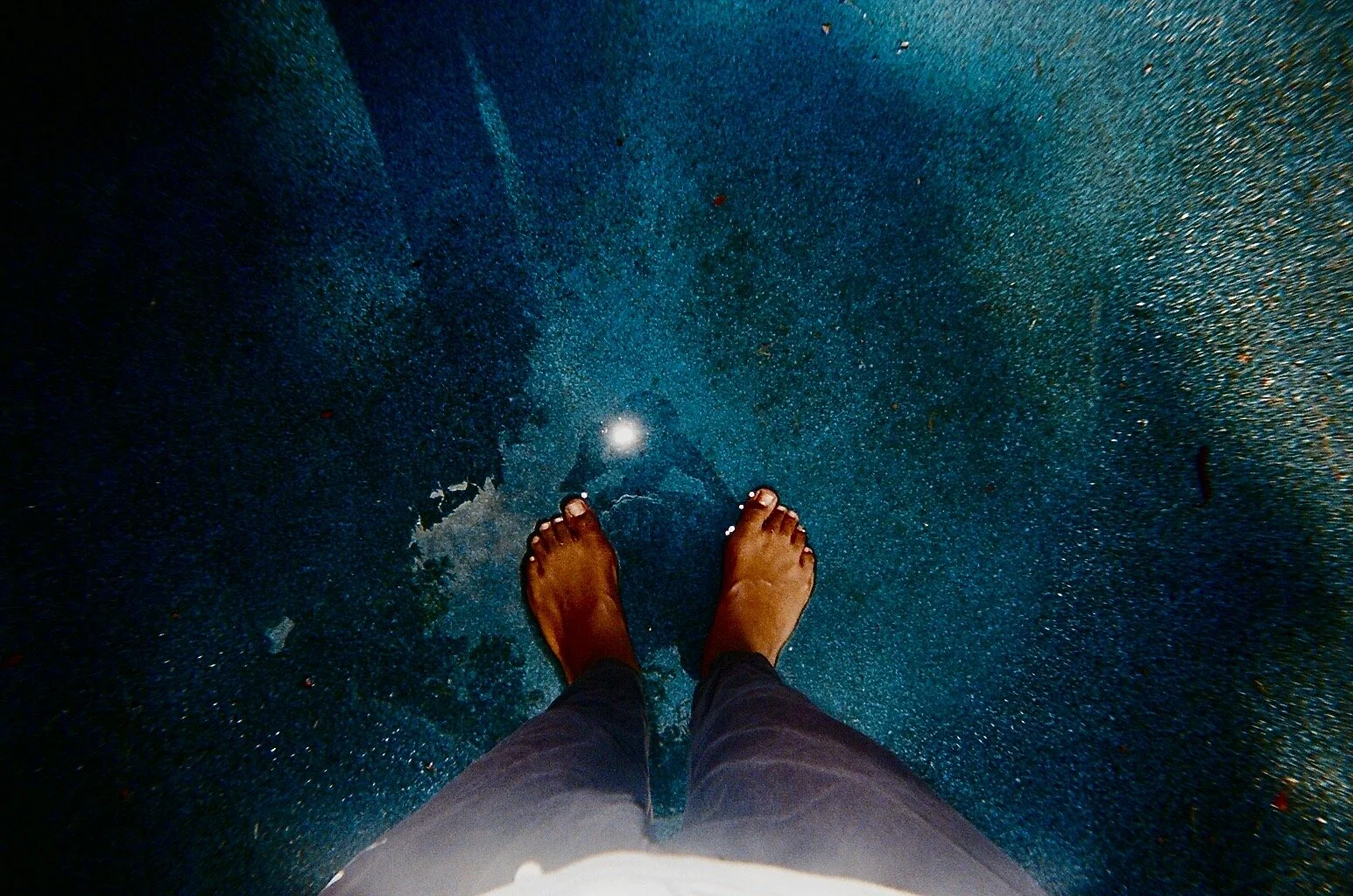 Person standing barefoot on a wet, shiny blue surface, possibly pavement, with feet near the center of the image and a bright reflection or light spot visible on the ground.
