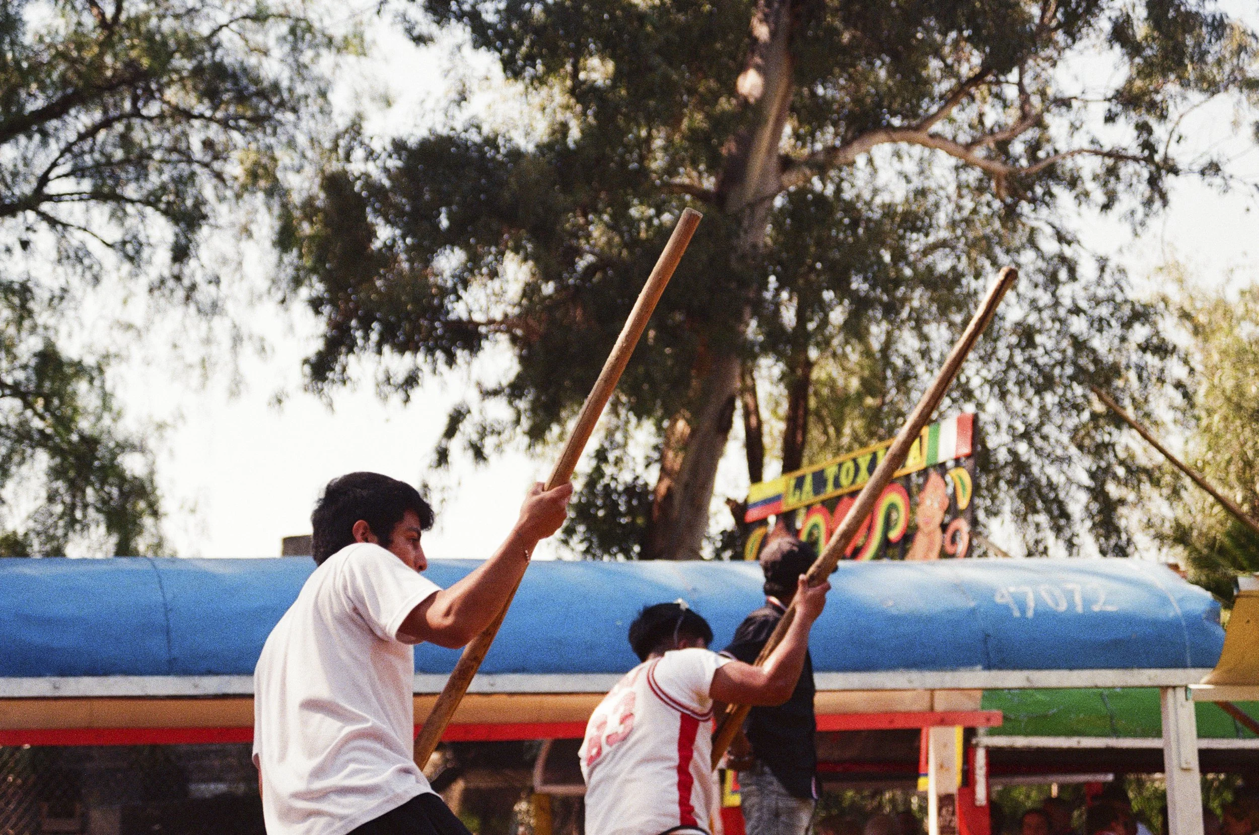 Group of young people holding wooden sticks at a fair or carnival, with colorful sign and trees in the background.