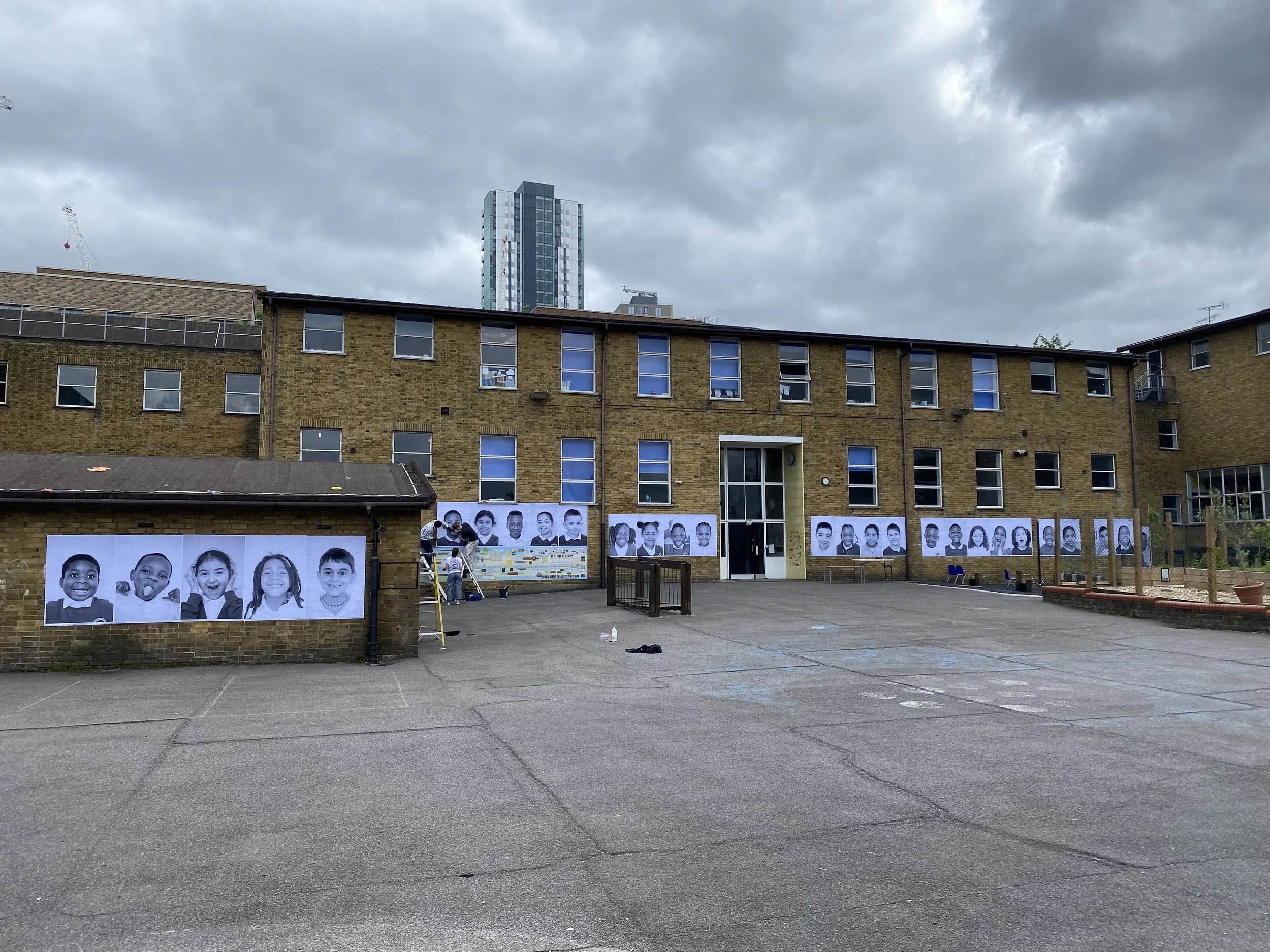 An outdoor courtyard area in front of a school with black and white photos of children on the wall, a ladder, and several adults working on a mural or display. There are tall residential buildings in the background under cloudy skies.