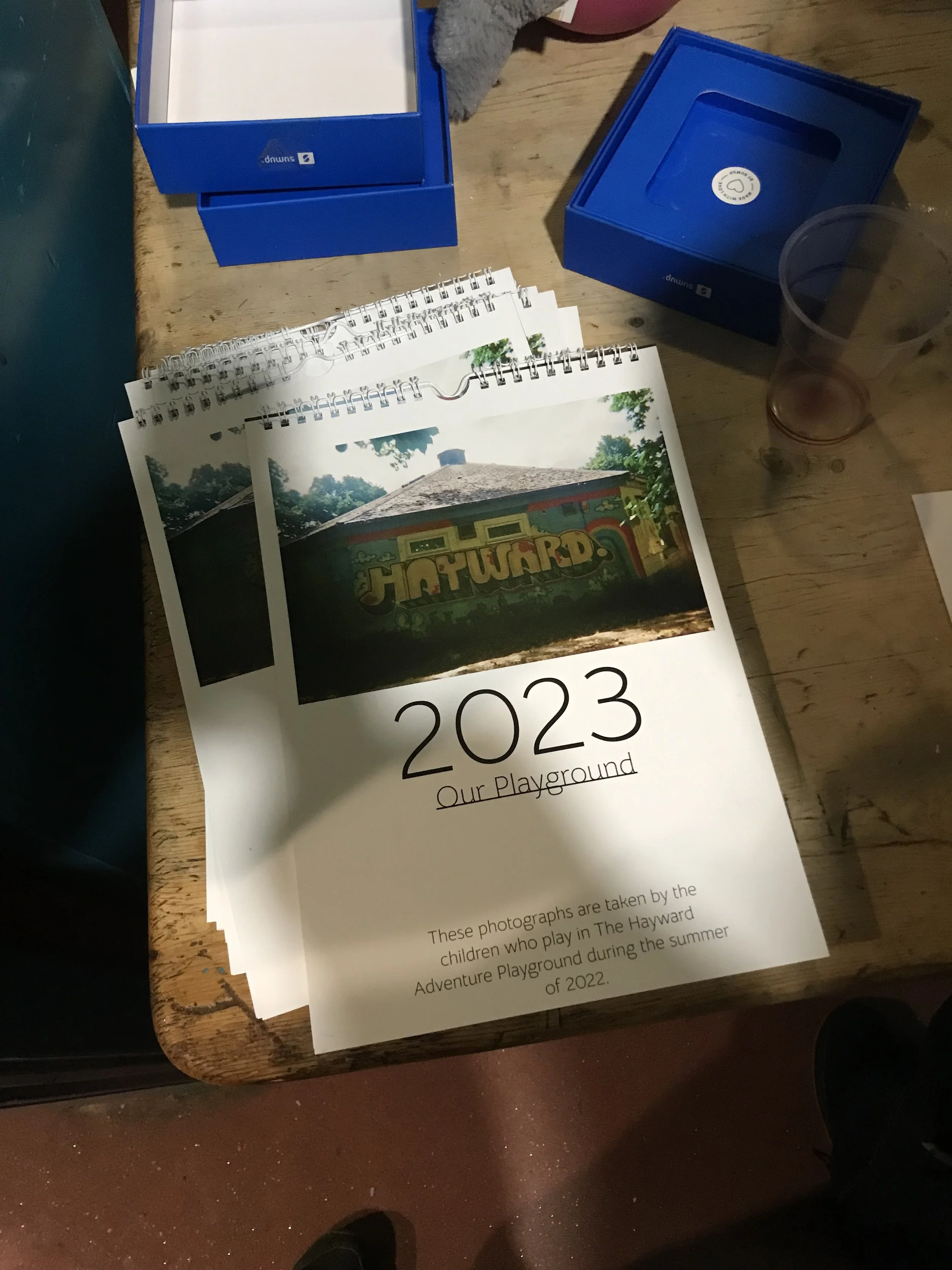 Photograph of a wooden table with blue boxes, photographs, and a white paper with the year 2023 and the words 'Our Playground' on it. The photograph appears to be part of a display or project related to children playing in a playground during summer.