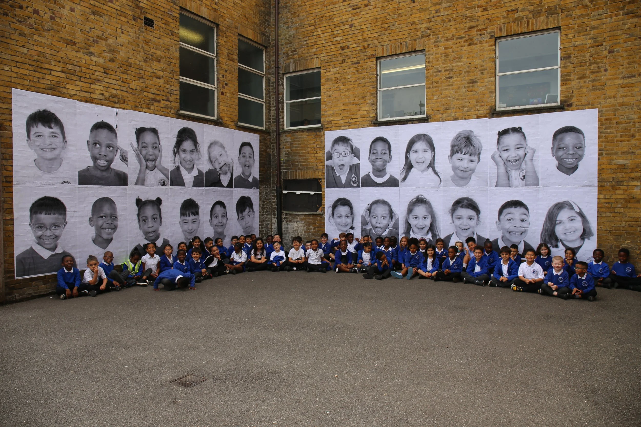 A large group of school children sitting on the ground in front of giant black-and-white portraits of children on a brick wall.