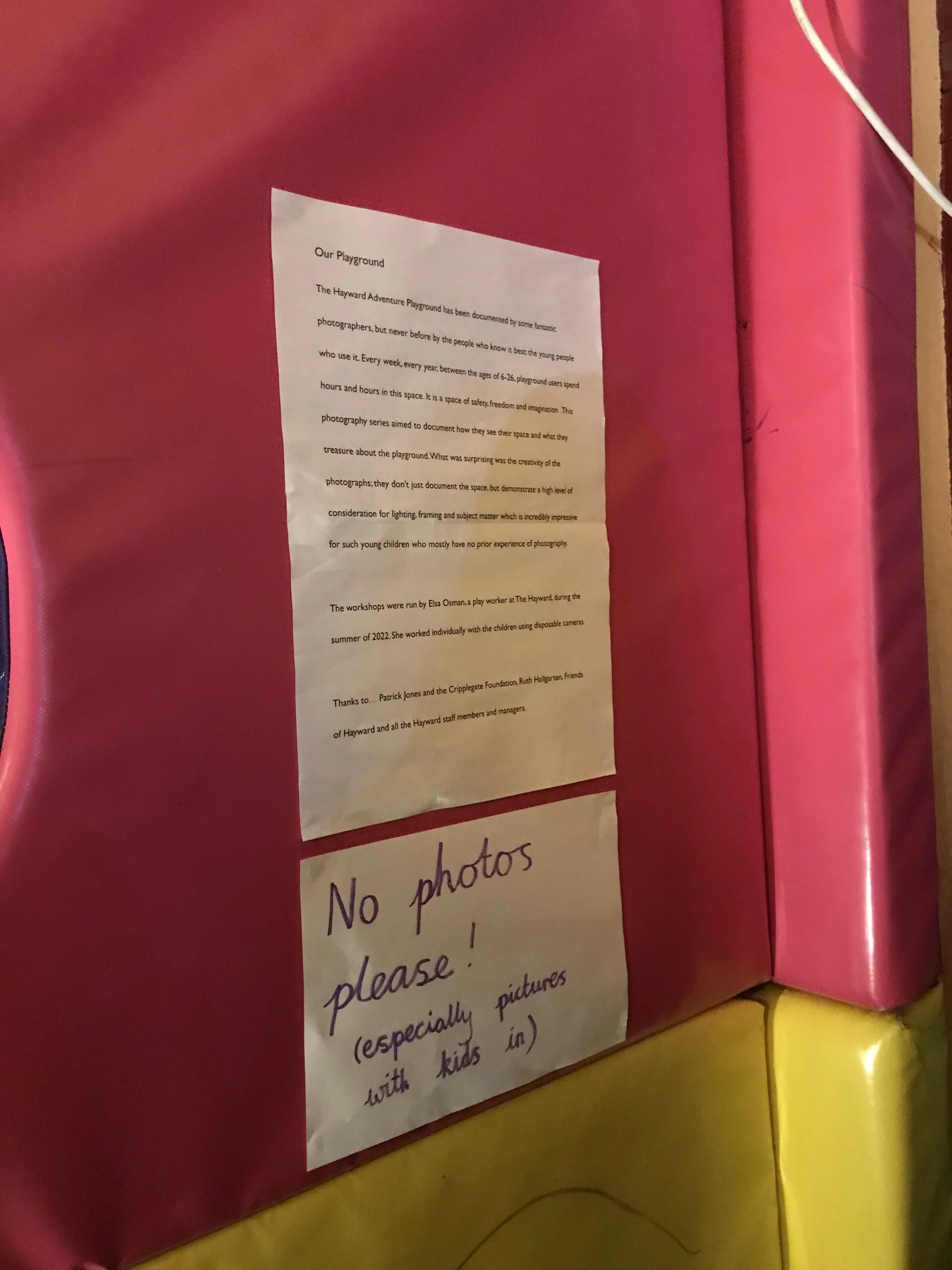A pink display board with two notices attached, one printed and one handwritten. The printed notice discusses a playground and photography, and the handwritten notice states 'No photos please!'