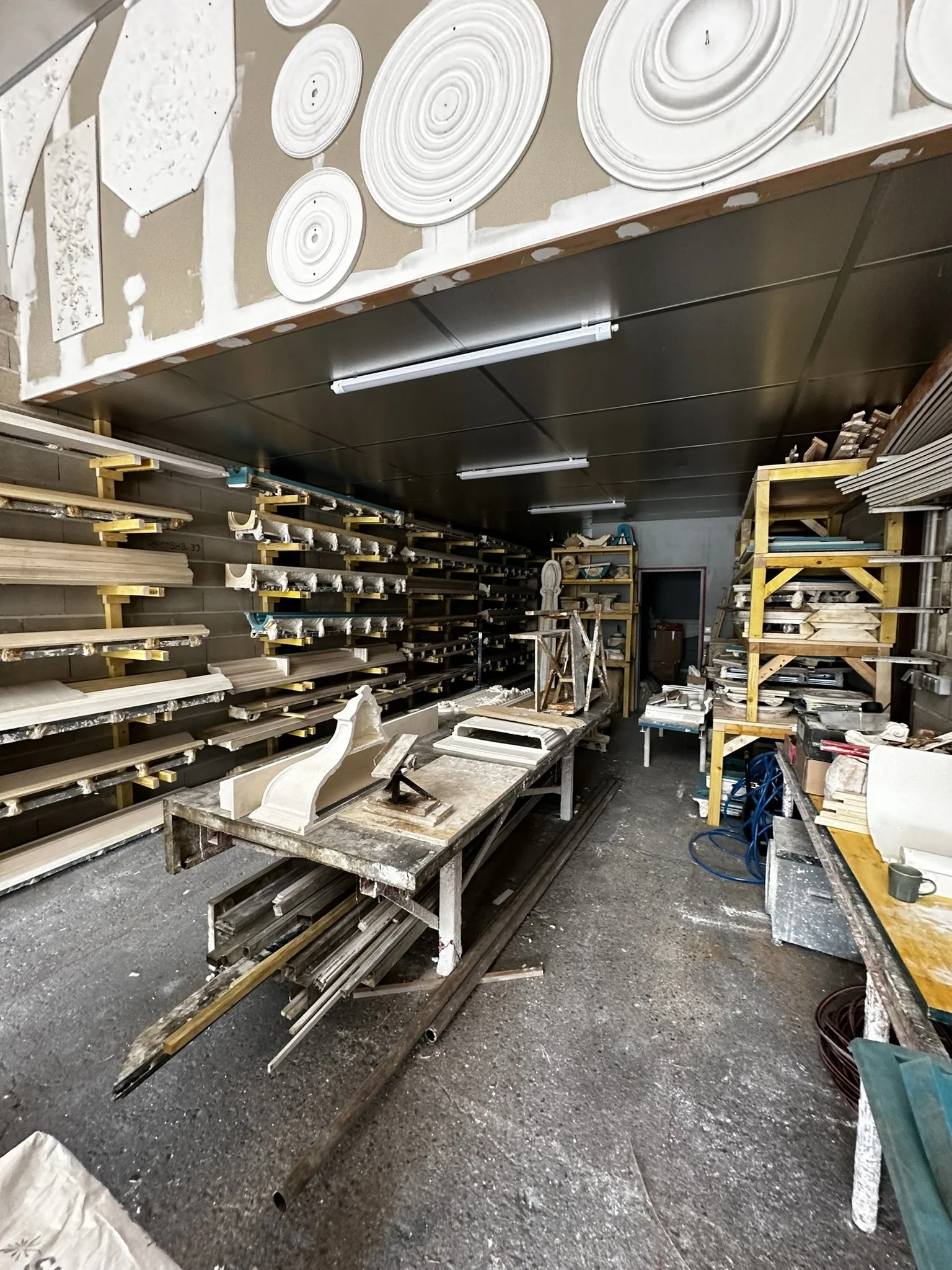 Une atelier de menuiserie avec des étagères remplies de moulures en bois, un bureau encombré d'outils et de morceaux de bois, murs partiellement peints, et un sol en béton sale.