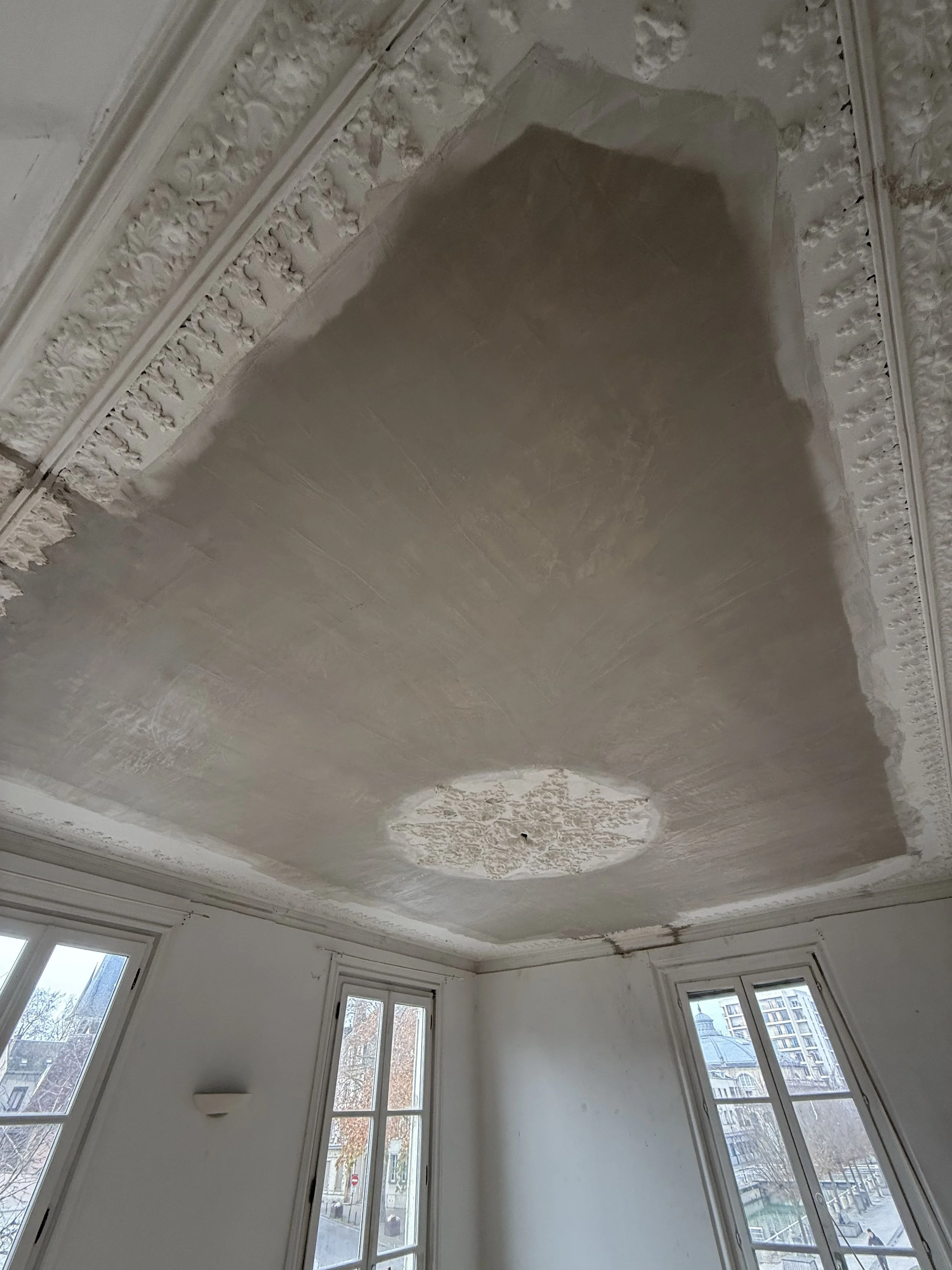 Vue intérieure d'une pièce avec grande fenêtre, plafond en cours de rénovation avec moulures ornementales et rosaces