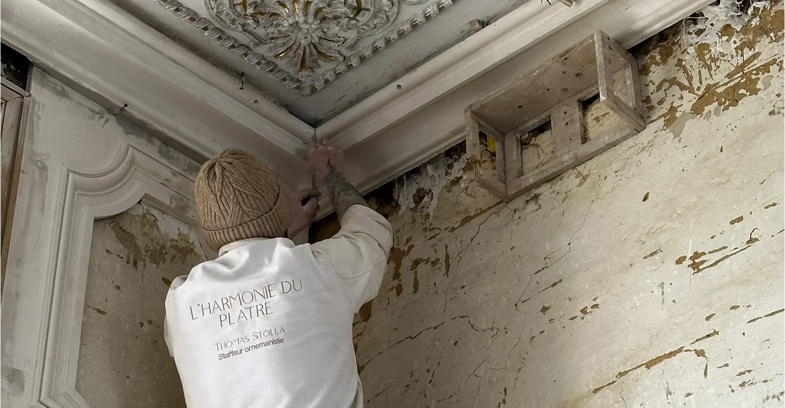 Un artisan en train de restaurer la décoration ornementale d'un plafond ancien, portant un sweat-shirt blanc avec l'inscription 'L'HARMONIE DU PLATRE' et une écharpe en laine, dans un espace avec des murs dégradés.