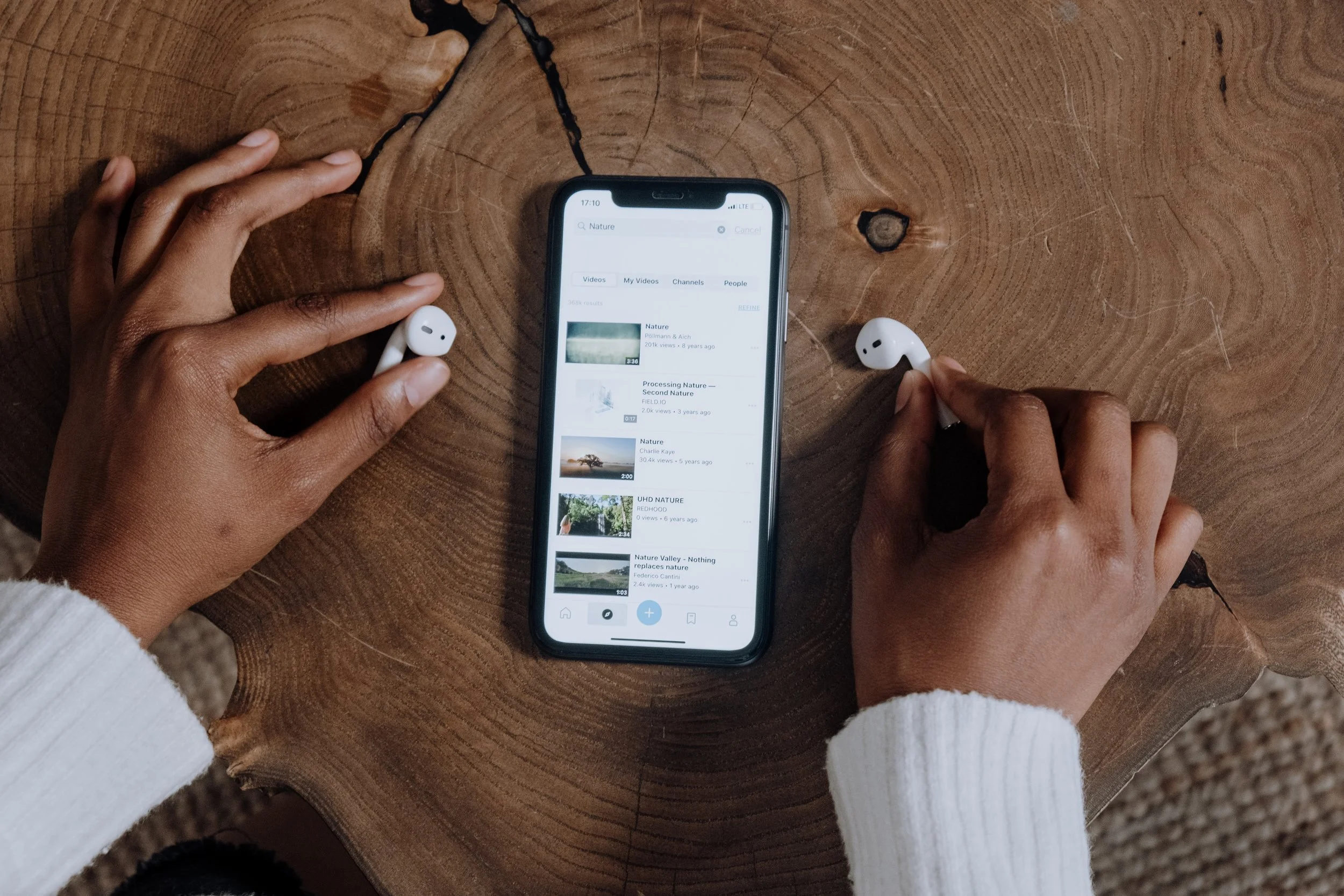Person holding wireless earbuds next to a smartphone displaying a YouTube search for nature videos, on a wooden table.