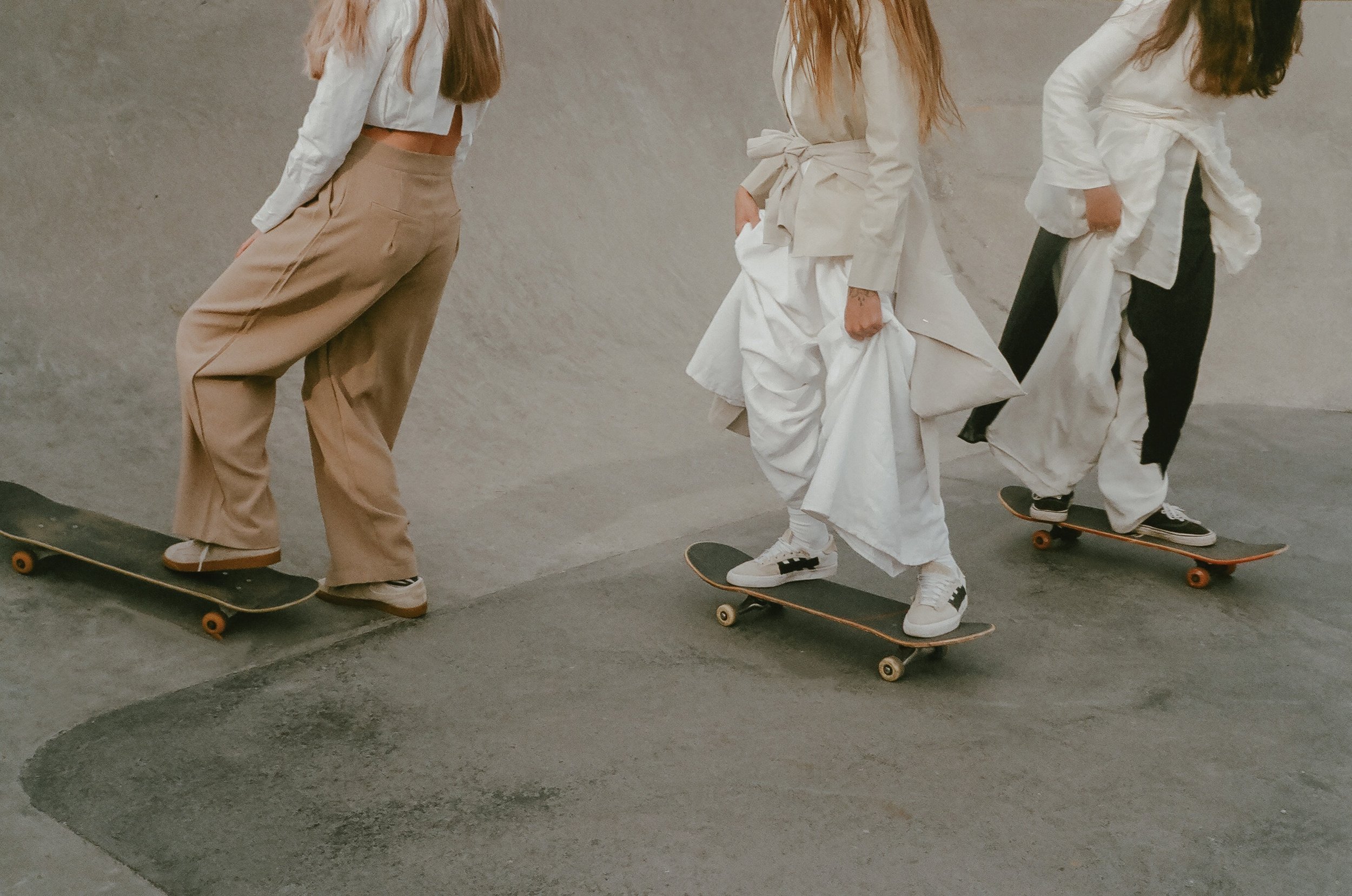 Three women skateboarding, wearing casual and oversized clothing in an indoor skate park.