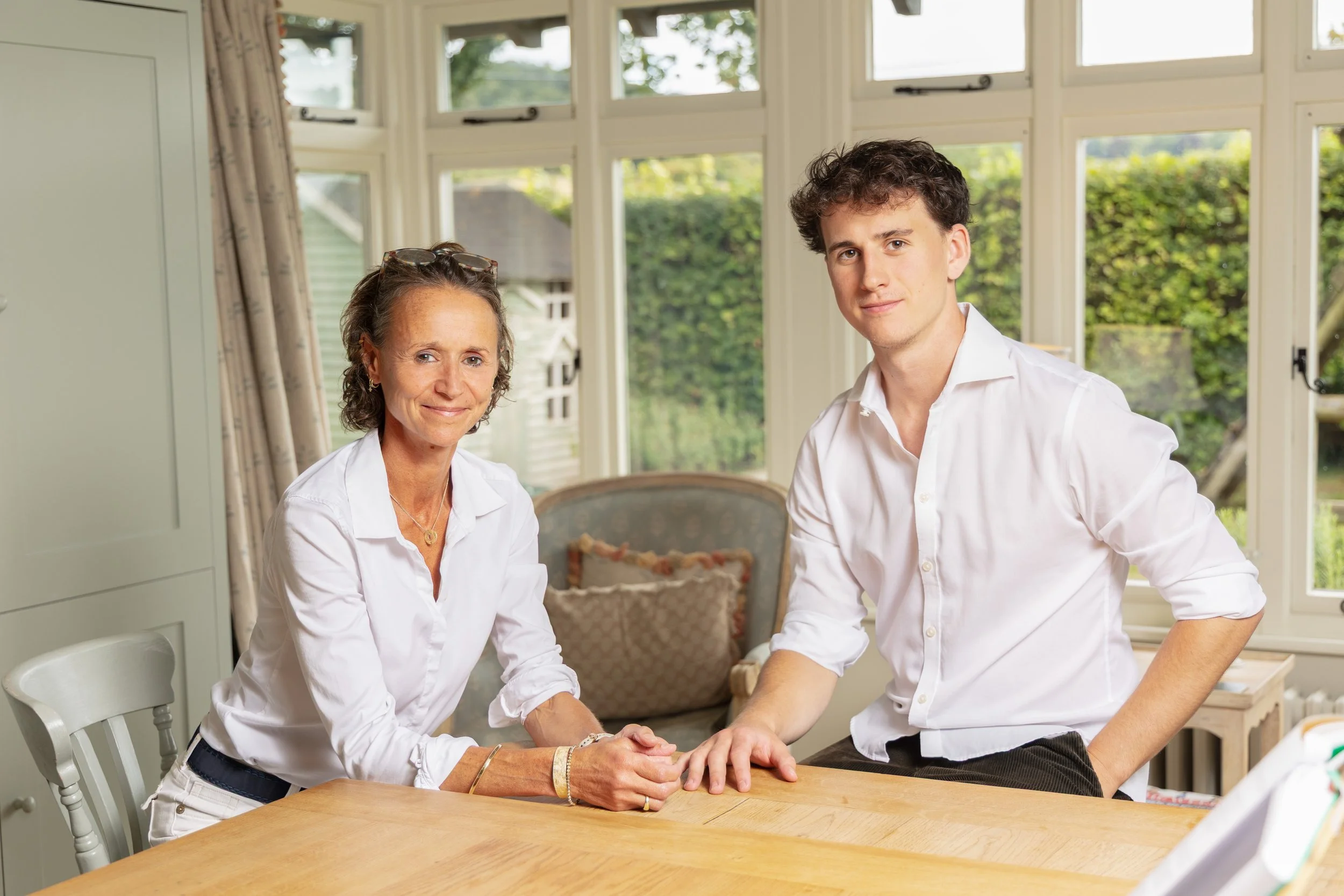A woman and a young man sitting at a wooden table in a bright room with large windows and green outdoor scenery in the background. The woman is smiling and wearing a white shirt, and the young man is resting his arm on the table, also dressed in a white shirt.