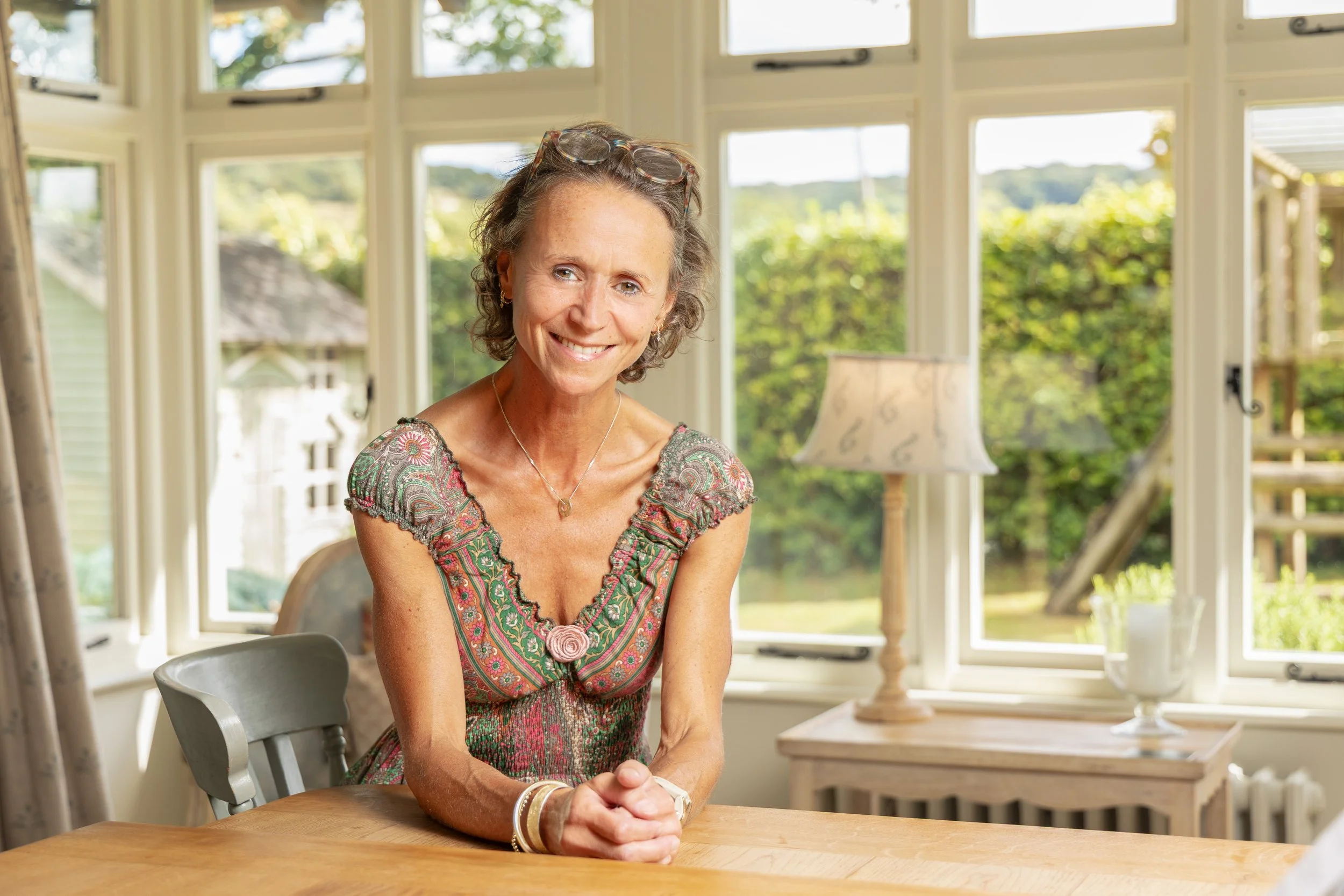 A smiling woman sits at a wooden table in a bright room with large windows, greenery outside, and a table lamp in the background.