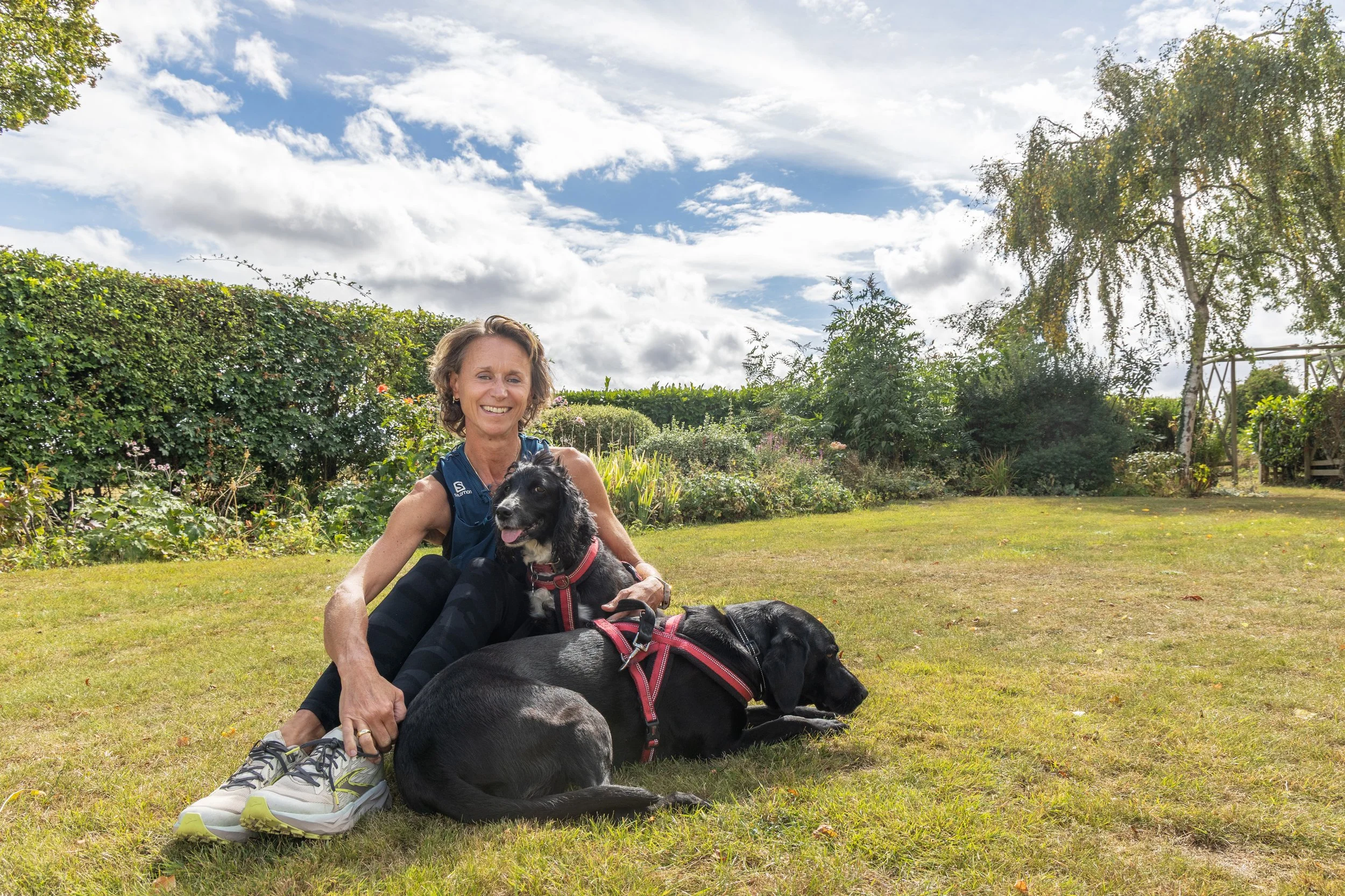 A woman sitting on the grass with two black dogs, smiling, in a garden with bushes, trees, and a partly cloudy sky.