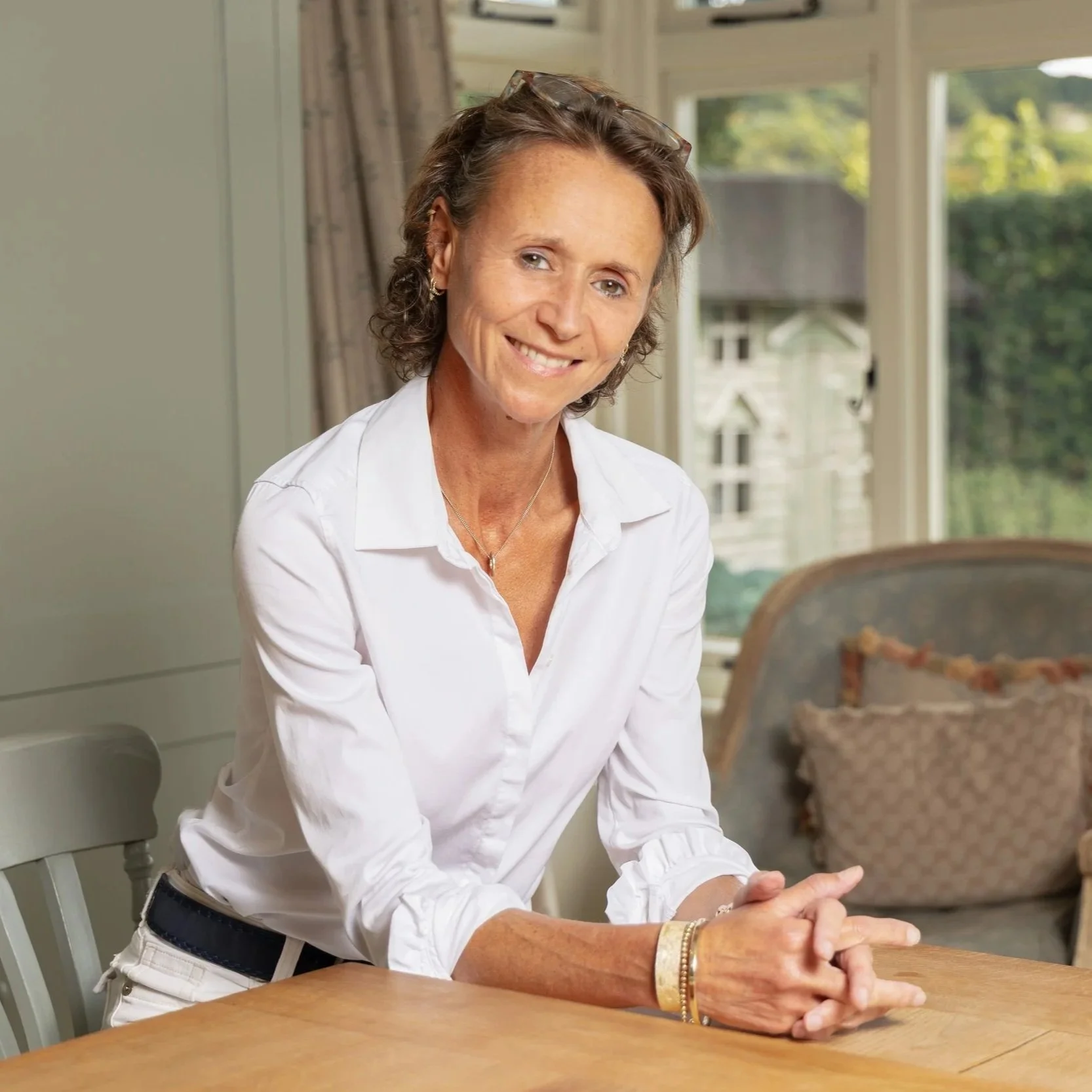 A smiling middle-aged woman with short curly brown hair, wearing a white button-up shirt, sitting at a wooden table inside a room with large windows showing greenery outside.