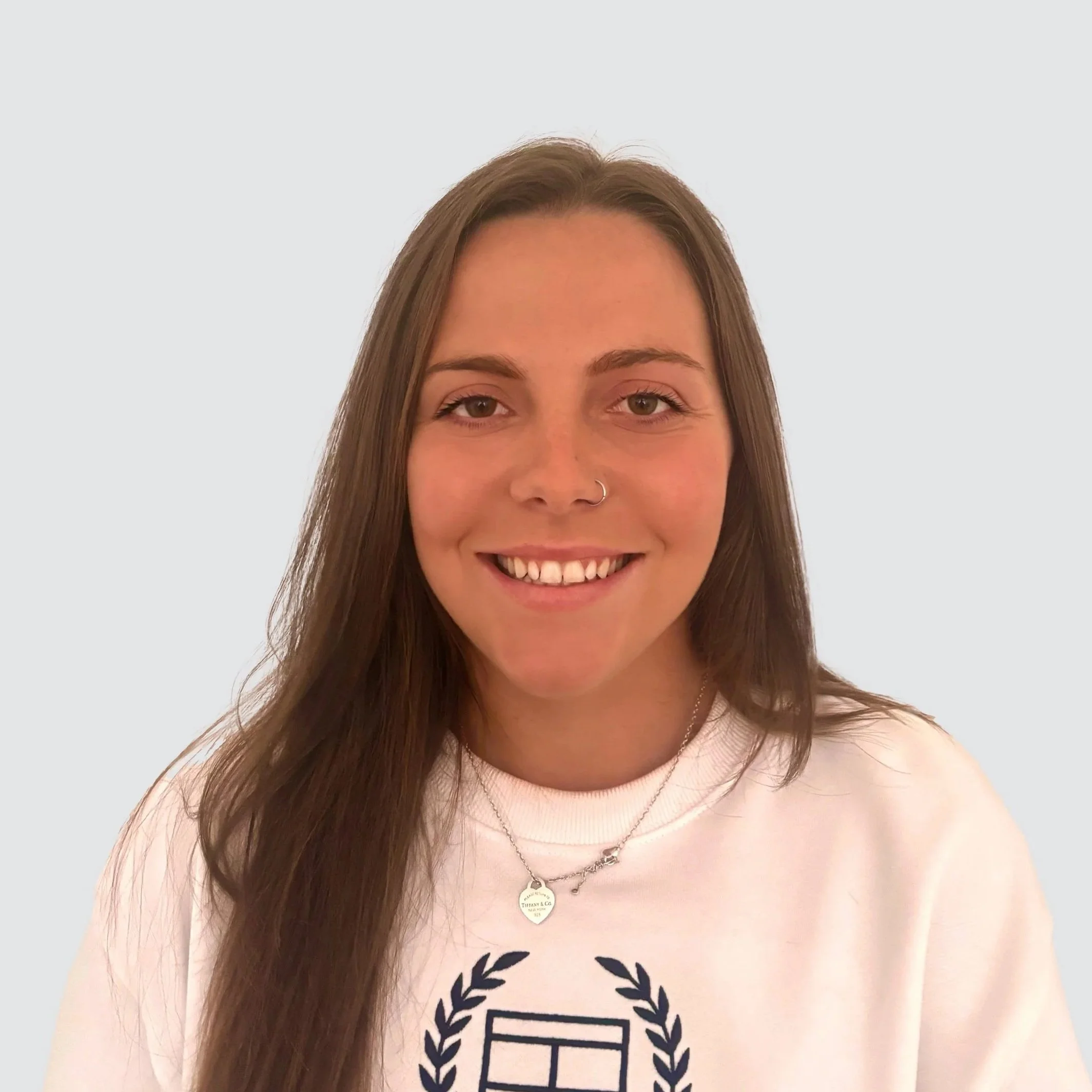 Smiling young woman with long brown hair, wearing a nose ring and layered necklaces, dressed in a white sweatshirt with a graphic design, posing against a plain light background.
