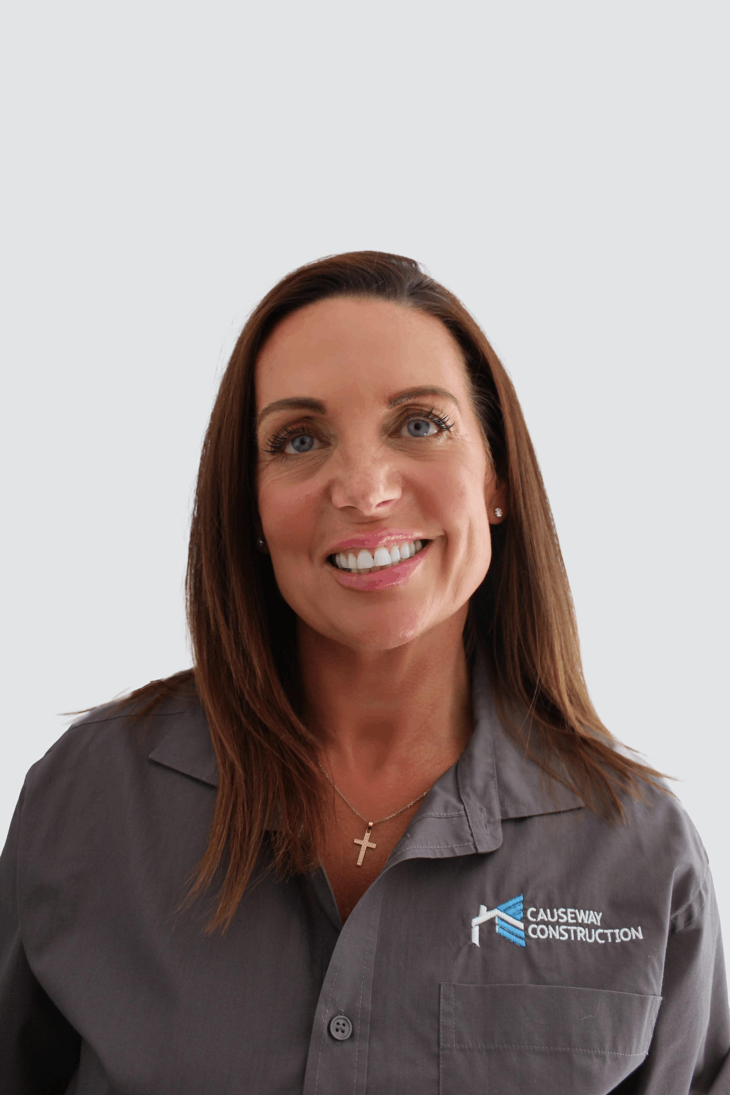 A woman with brown hair and blue eyes smiling, wearing a gray collared shirt with a 'Causeway Construction' logo and a gold cross necklace, against a plain white background.