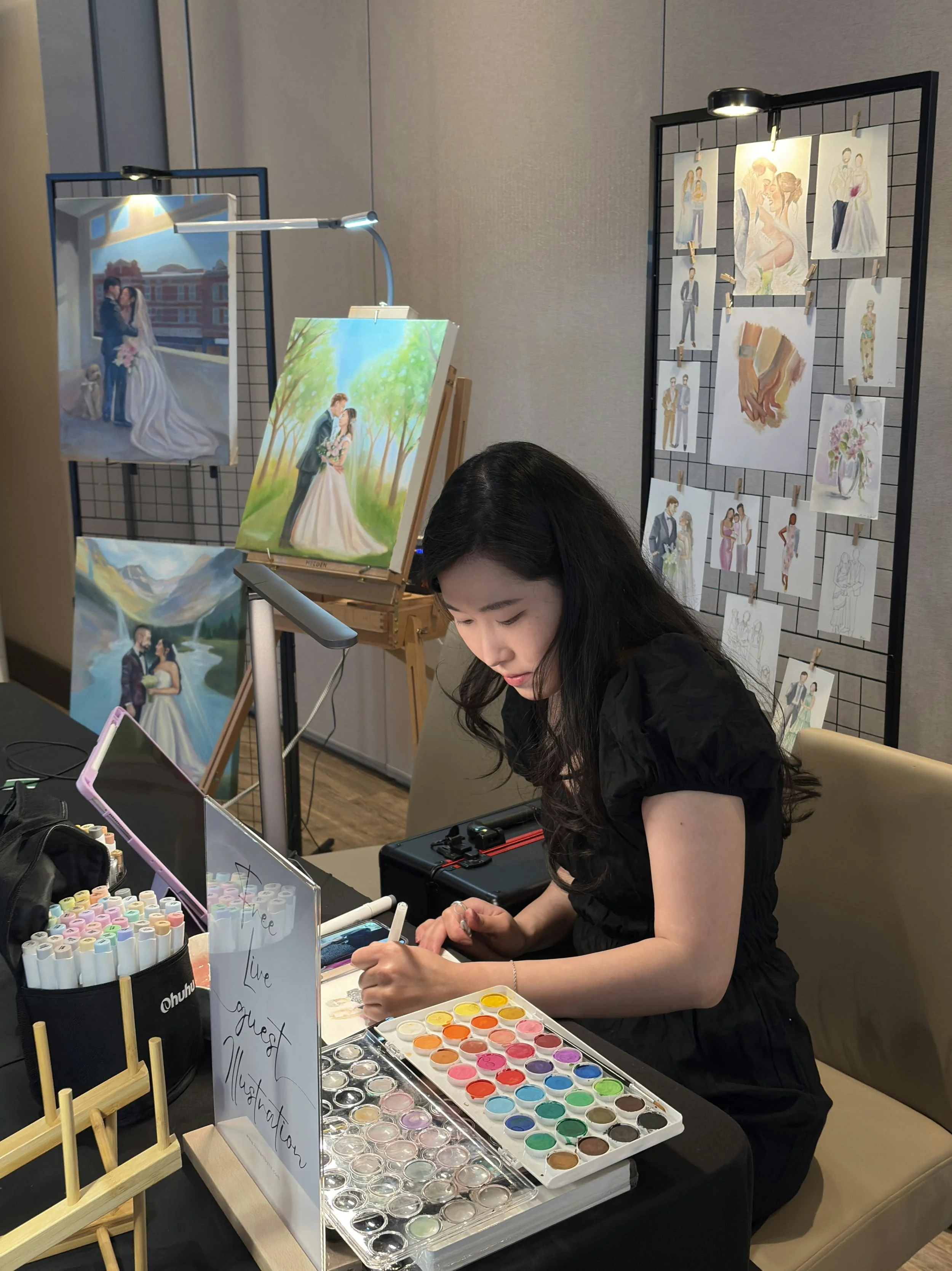 Liz working at an art table with watercolor paints, markers, and art supplies. Behind her, on display boards, are watercolor paintings of wedding scenes and fashion illustrations.