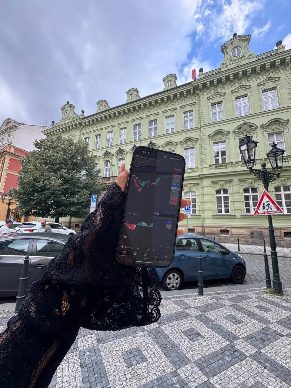 Person holding a smartphone displaying stock market charts, outdoors in front of a green building with ornate architecture, cars parked along the street, cloudy sky overhead.