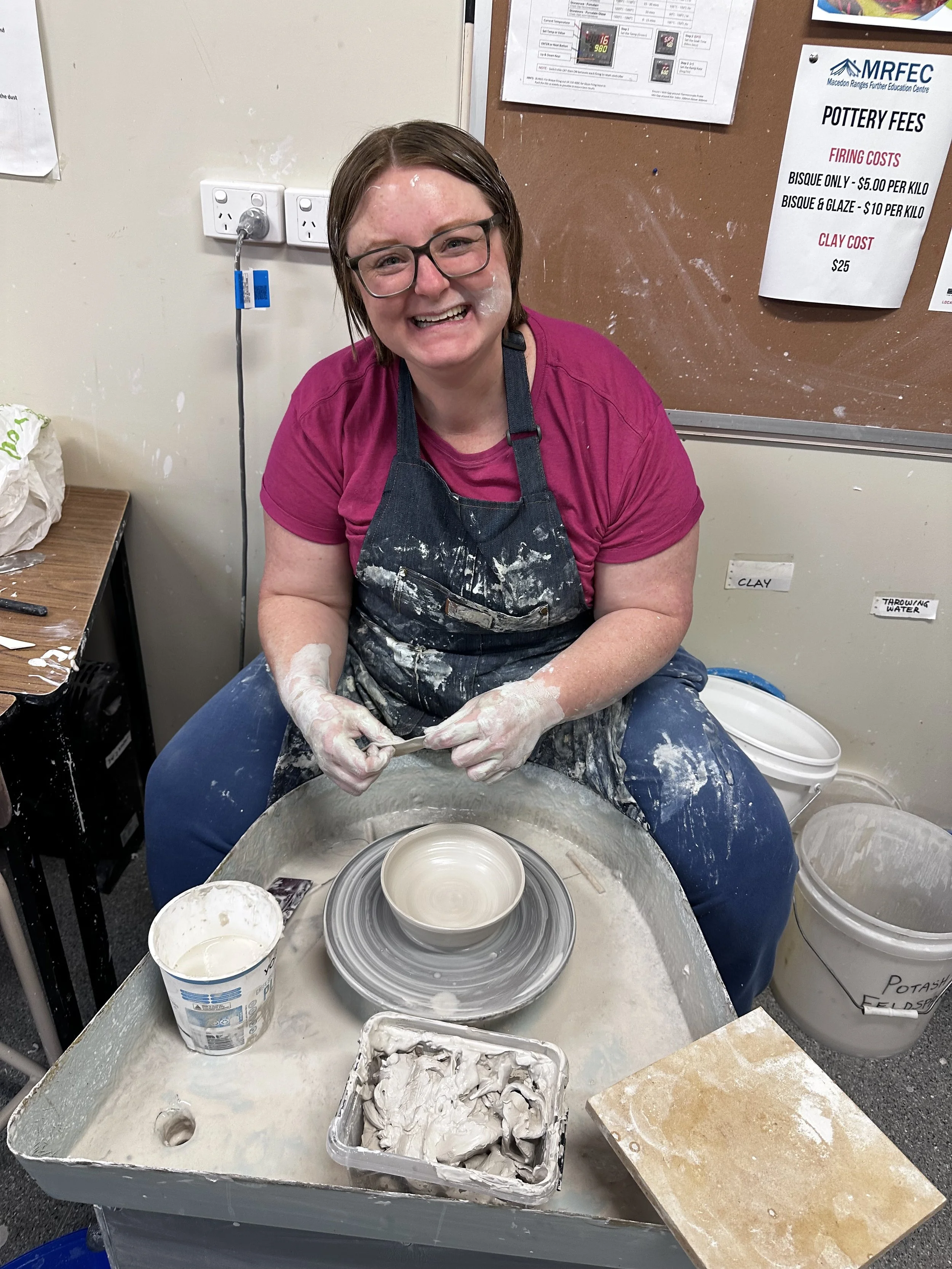 A smiling woman with glasses and short brown hair in a pottery studio, wearing a pink shirt and a dark apron, working on a pottery wheel with a ceramic piece. There are buckets of clay and plaster near her, along with a corkboard and posters with pottery fees and instructions in the background.
