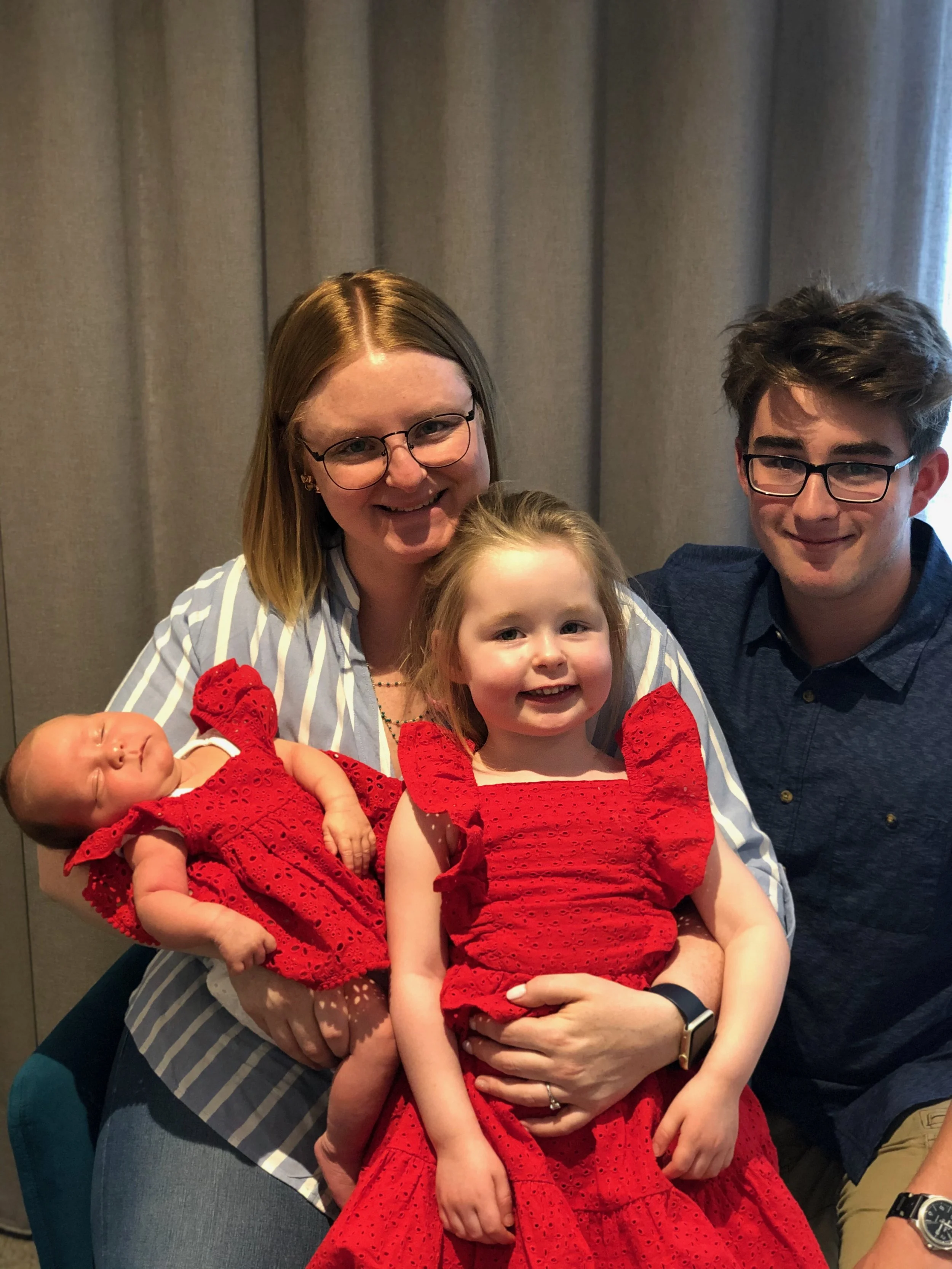 A smiling woman holding a sleeping baby dressed in red lace with a young girl in matching red lace dress, and a young man smiling beside them, standing in front of gray curtains.