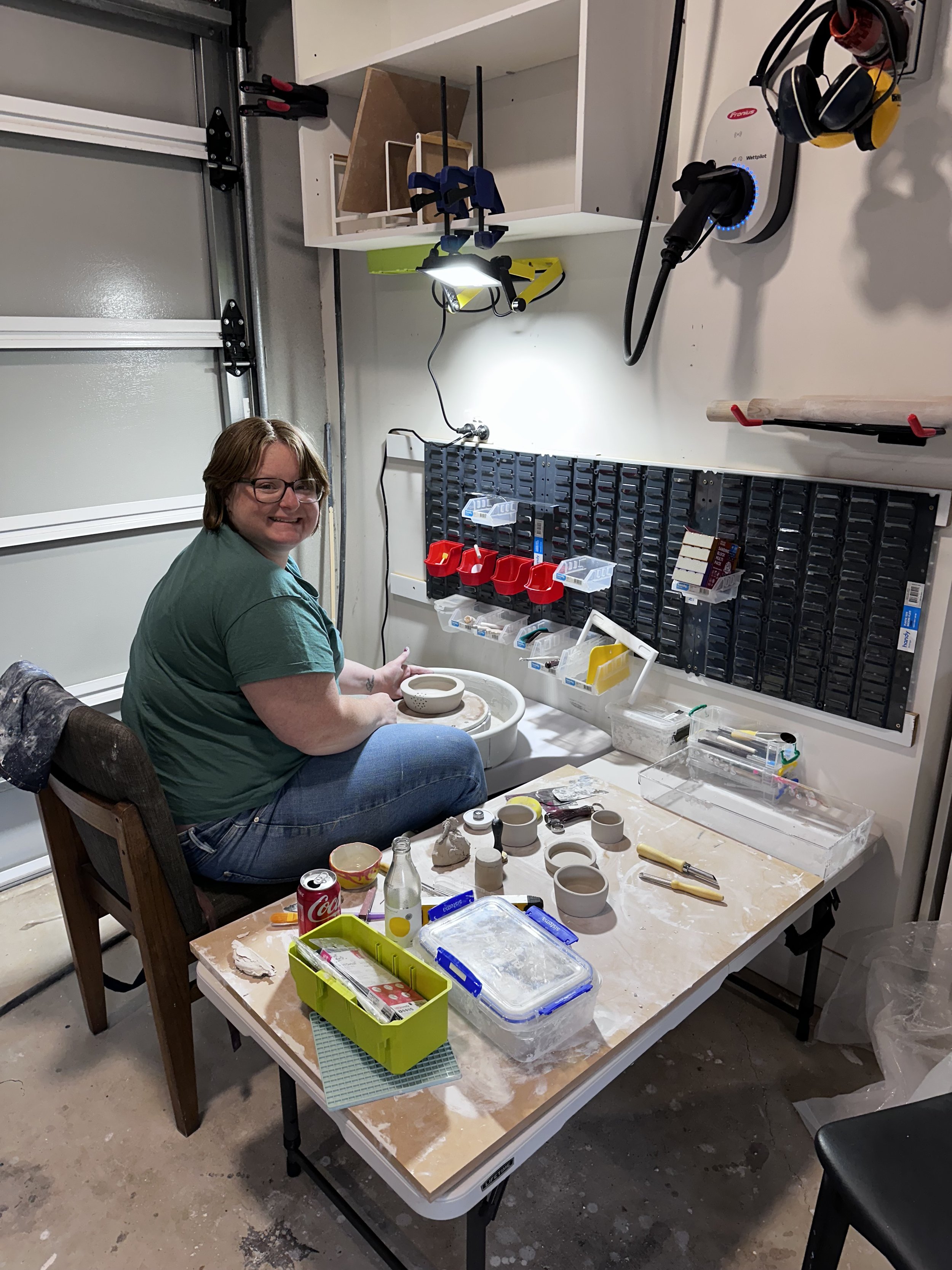 A person working on a pottery wheel in a garage studio, surrounded by tools, pottery, and storage containers.