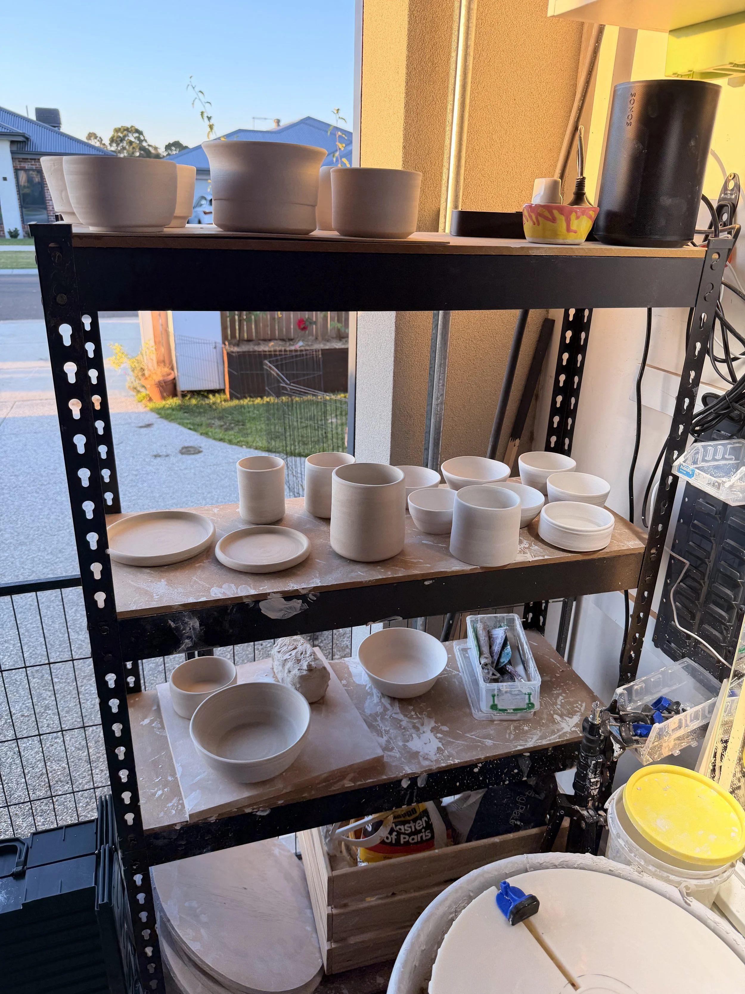A black metal shelving unit with three wooden shelves, holding unfinished ceramic pottery, including bowls, cups, and plates, in a workshop with tools, a window, and a view of a yard outside.