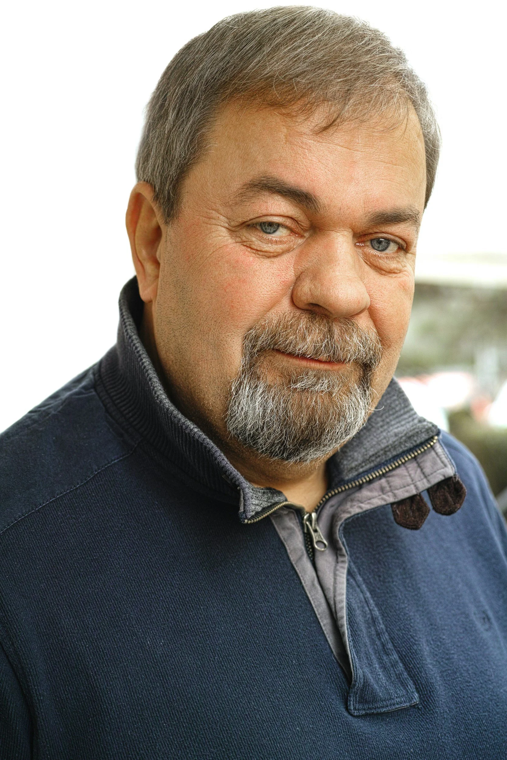 Close-up of a middle-aged man with gray hair, a beard, wearing a navy blue jacket with a gray zip-up collar, looking slightly to the right with a slight smile.
