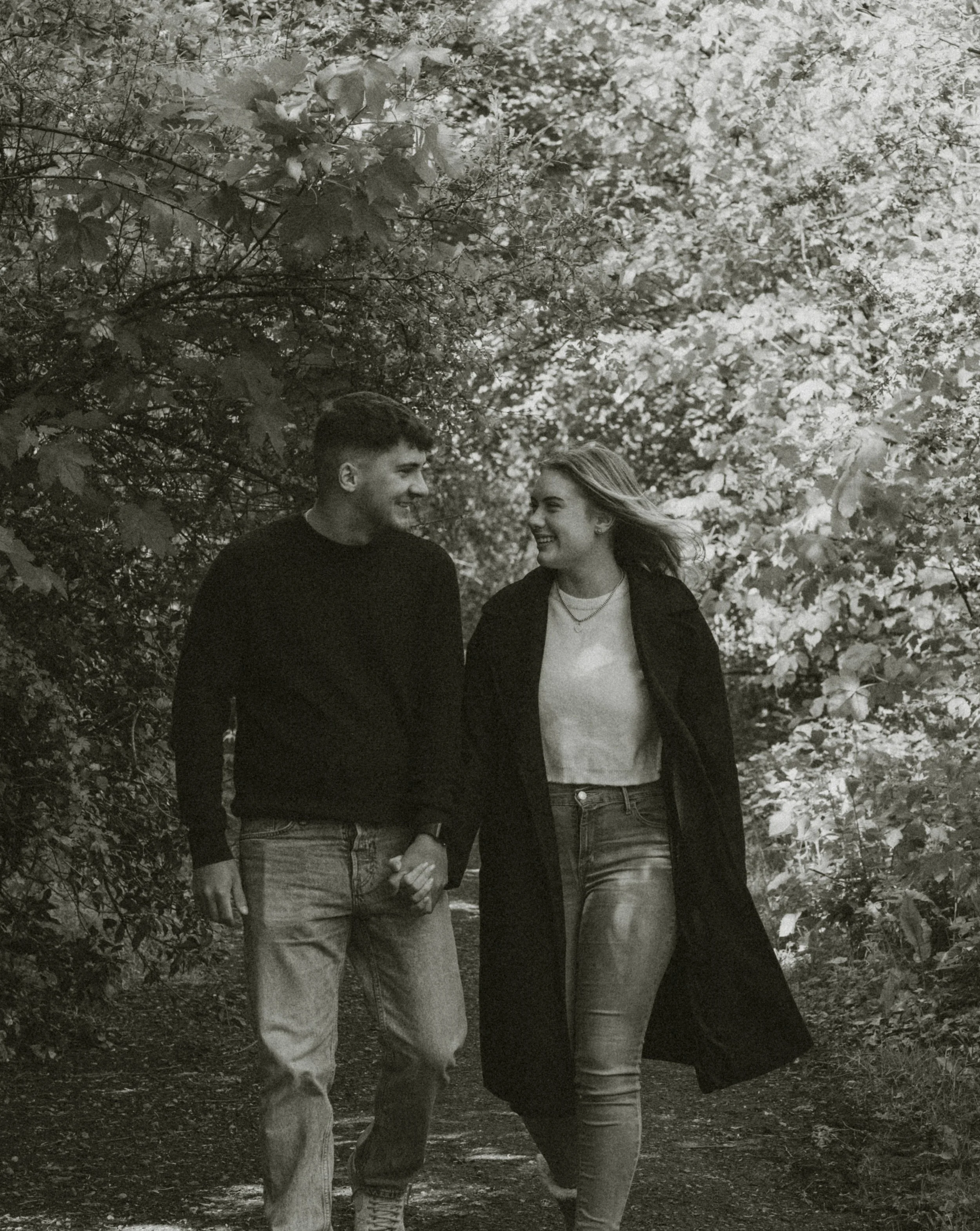 A black and white photo of a young man and woman walking together outdoors, surrounded by trees and foliage, smiling at each other.