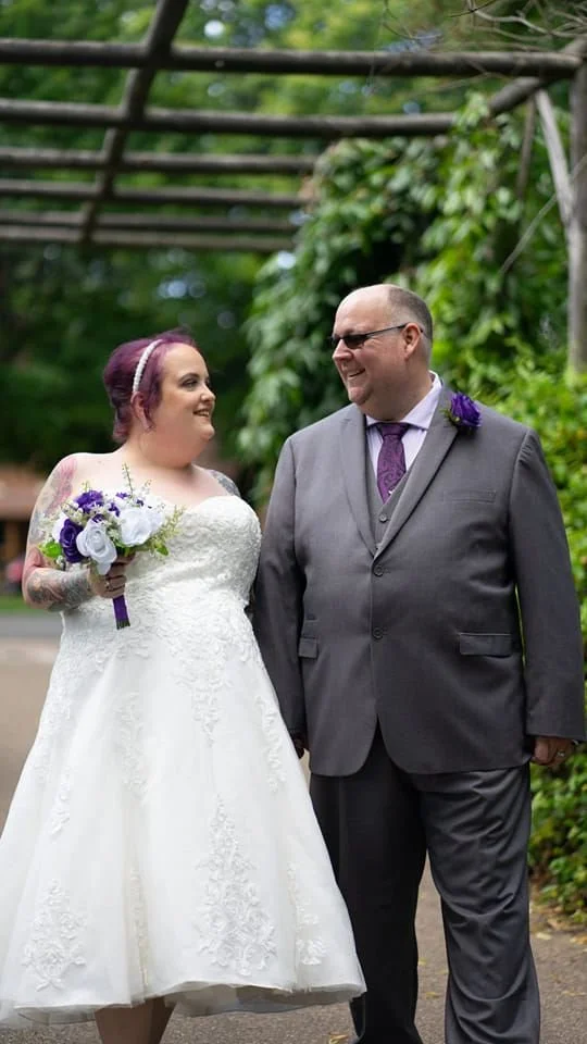 Bride and groom smiling at each other at a wedding outdoors, holding hands. The bride wears a white lace wedding gown with lace sleeves and a veil, holding a bouquet of purple and white flowers. The groom wears a gray suit with a purple tie and bouto
