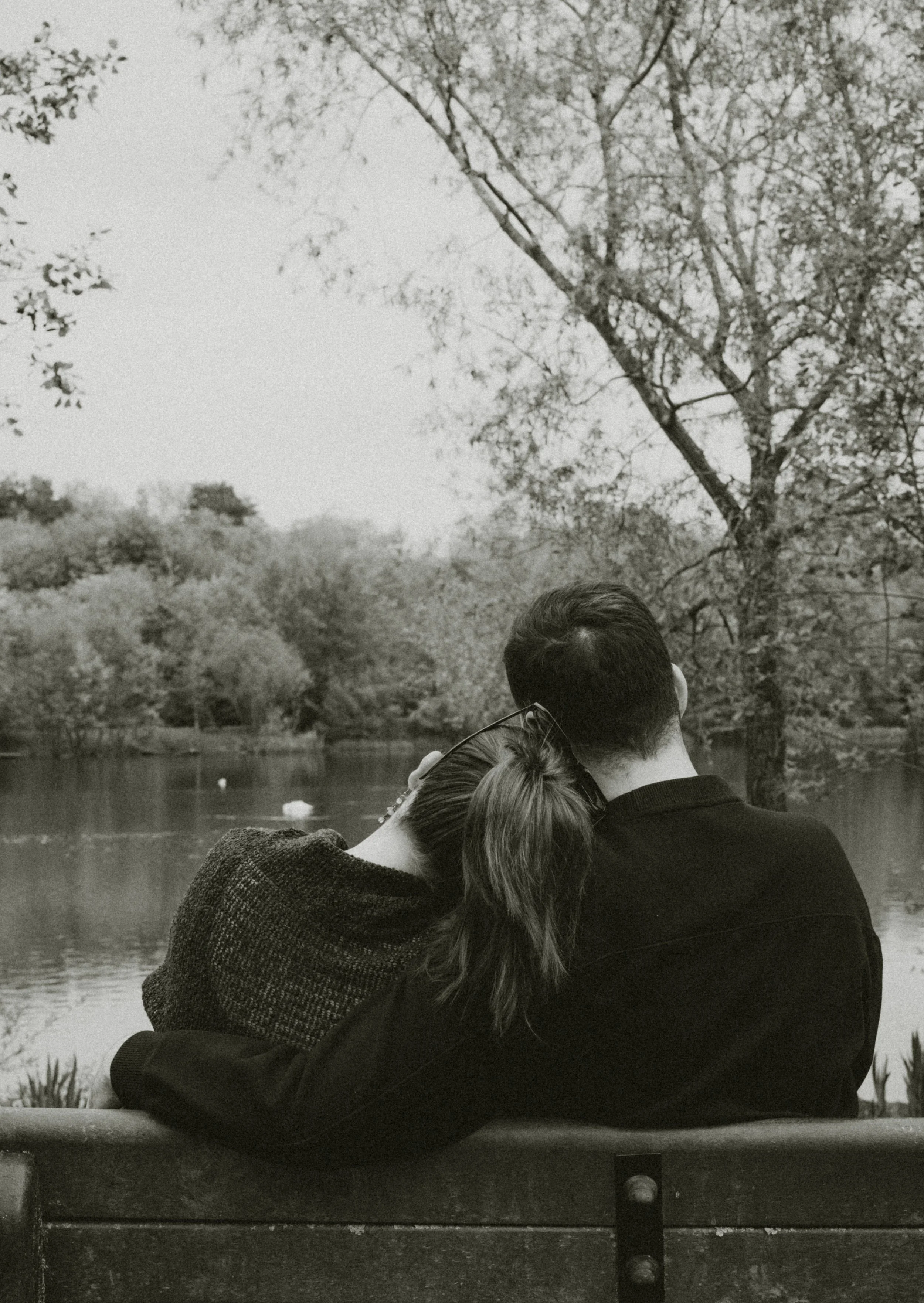 Two people are sitting on a park bench near a body of water with trees in the background. The woman is resting her head on the man's shoulder, and both are facing away from the camera.