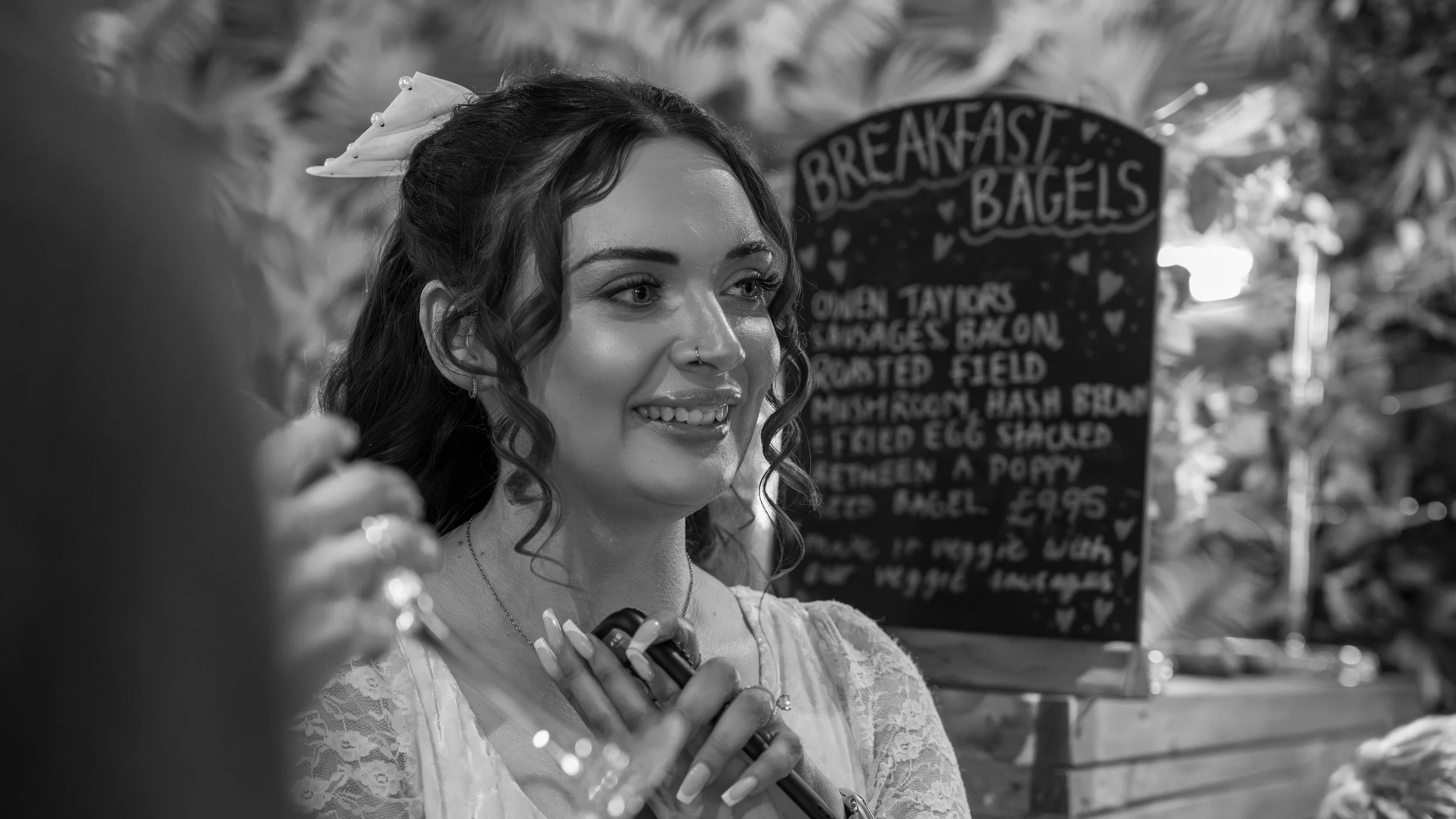A woman with dark, curly hair and a nose ring smiling and holding her hand over her chest at a market stall with a sign advertising breakfast bagels in the background.