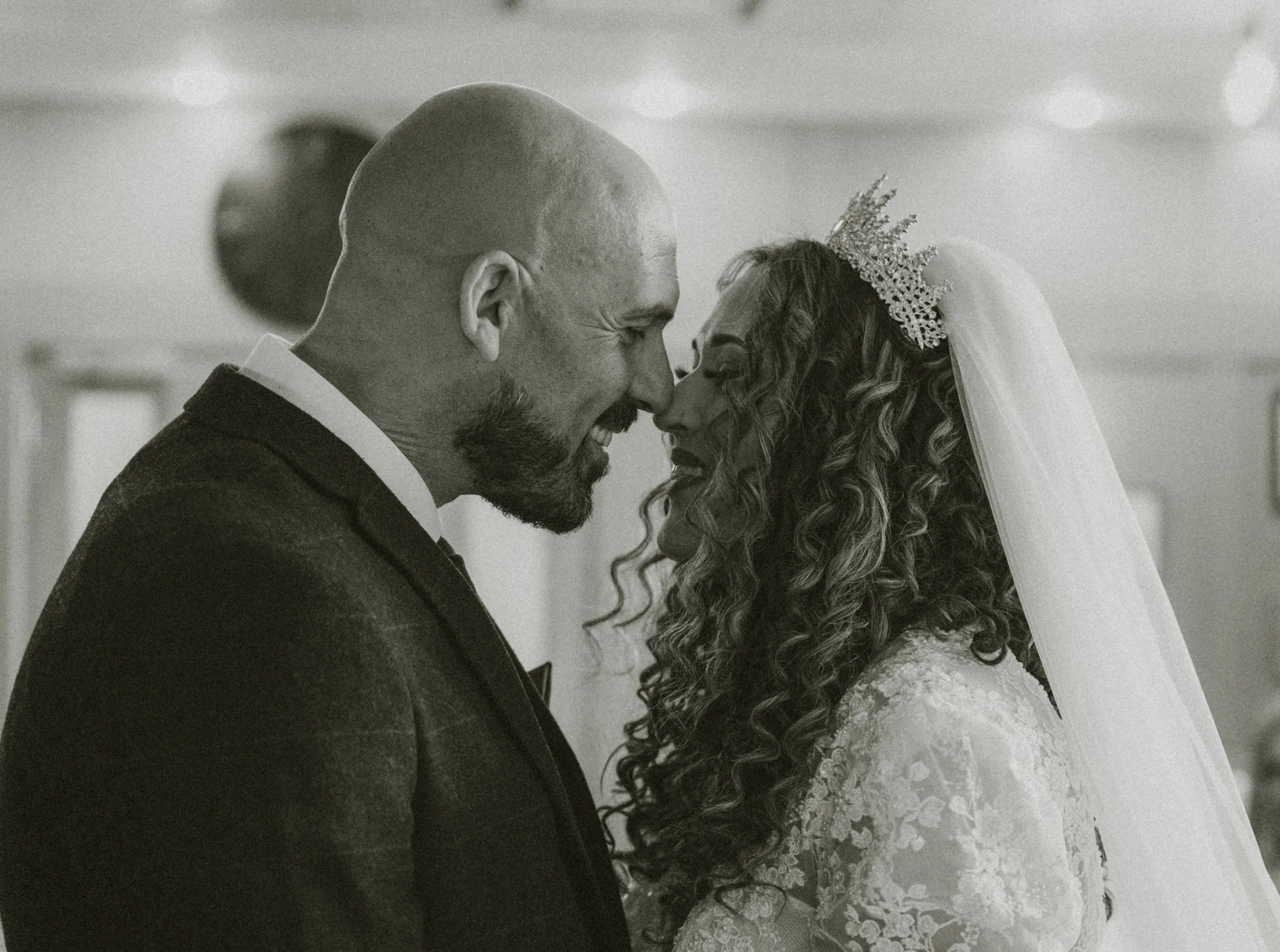A black and white photo of a bride and groom smiling and touching foreheads. The groom has a beard and is bald, wearing a dark suit. The bride has long, curly hair, a veil, and a decorative headpiece, wearing a lace wedding dress.