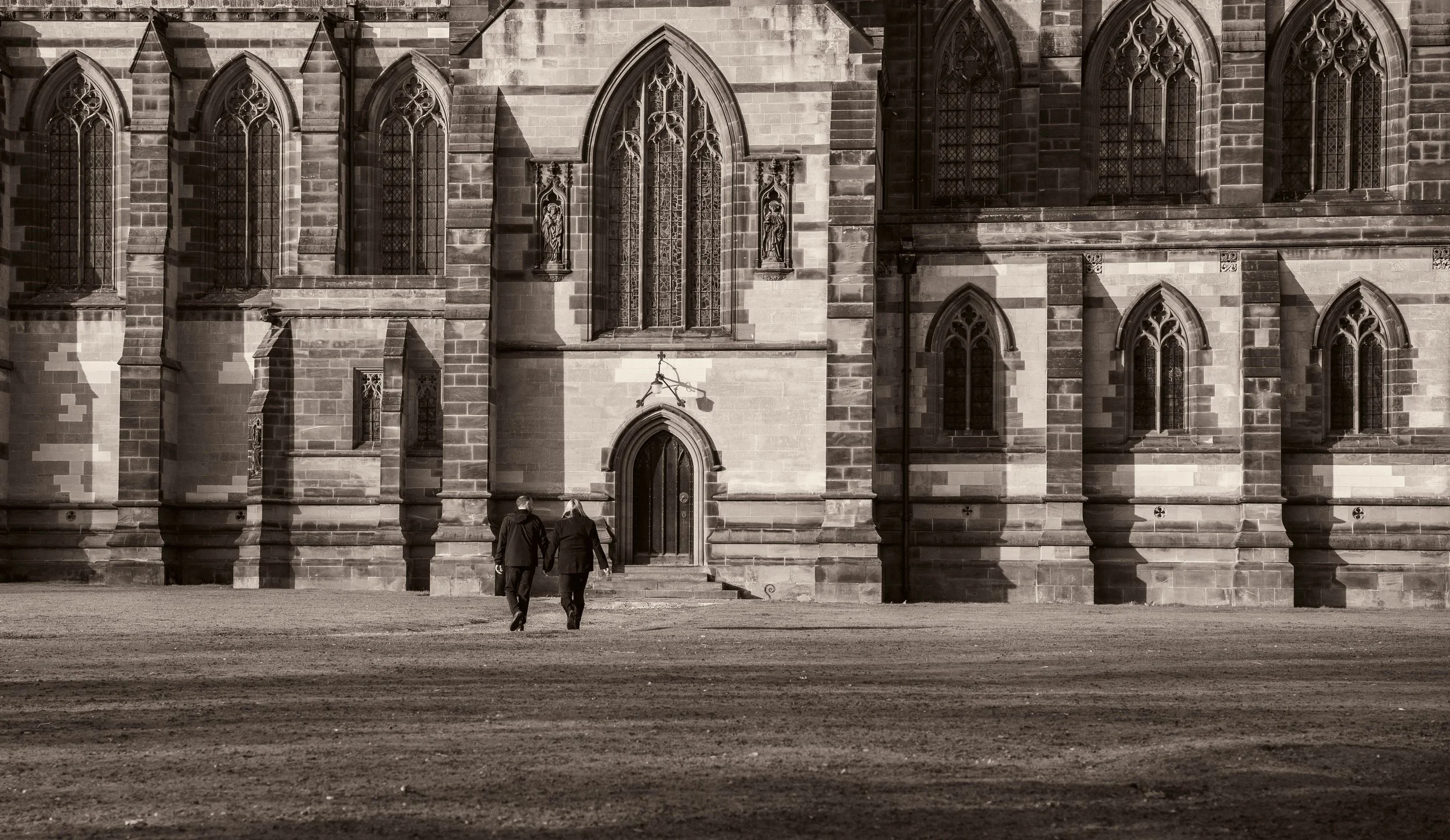 A black-and-white photo of a Gothic-style church with large arched windows, intricate stained glass, and stone statues. Two people are walking toward the entrance, holding hands.