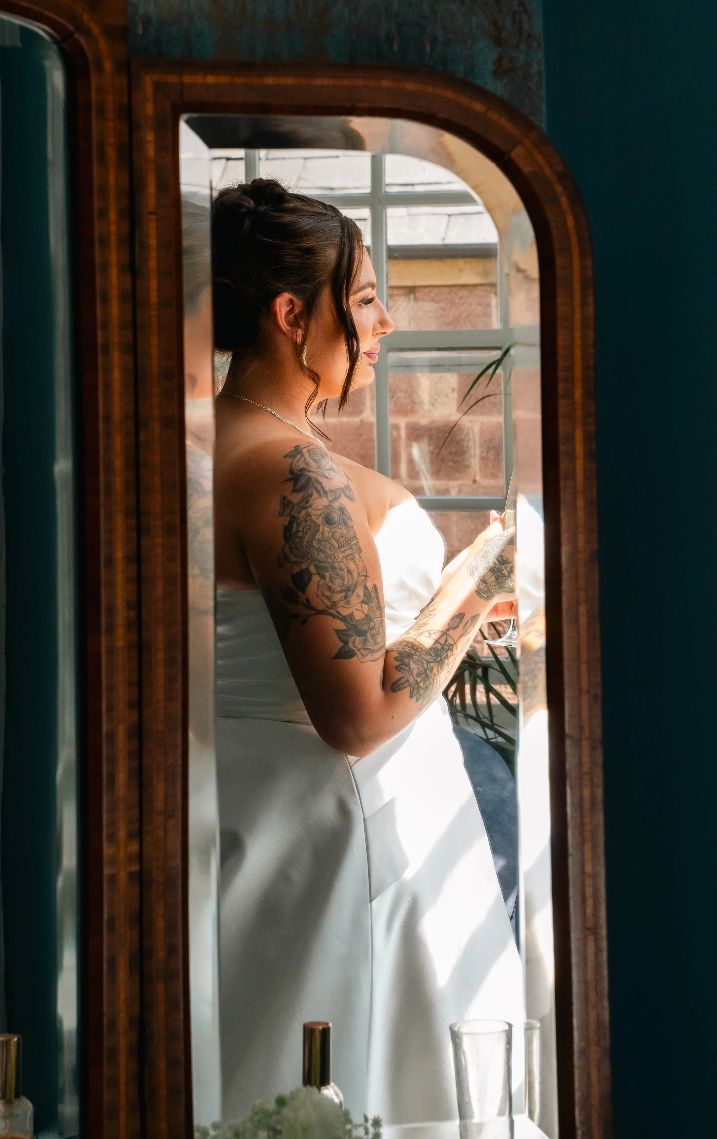 Woman in wedding dress with tattoos, looking at her reflection in a mirror while holding a glass, natural light coming through a window.