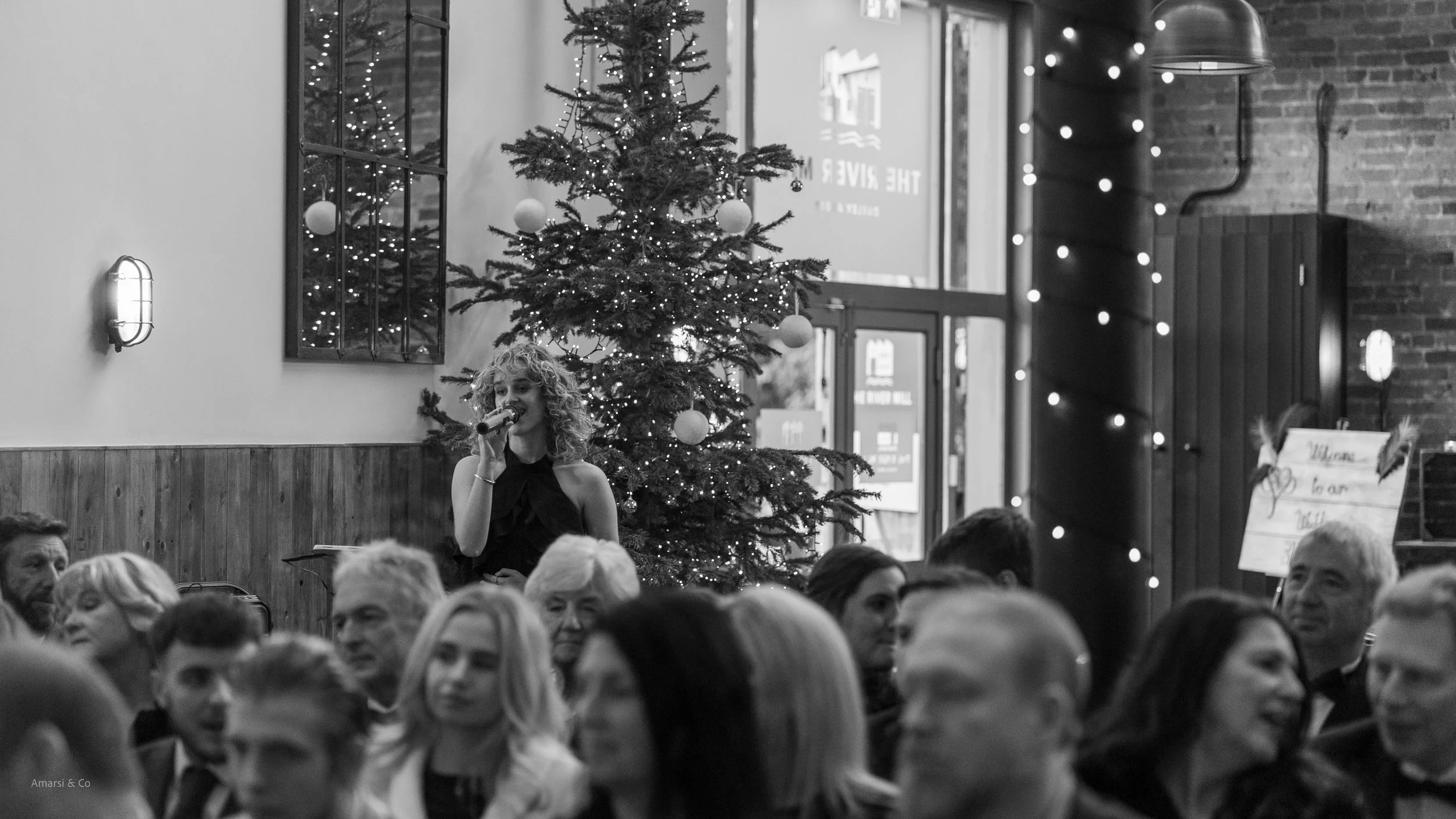 A black and white photo of a woman singing into a microphone during a festive gathering with a Christmas tree decorated with ornaments and lights in the background. People are seated in the foreground, and the setting is the river mill, derby