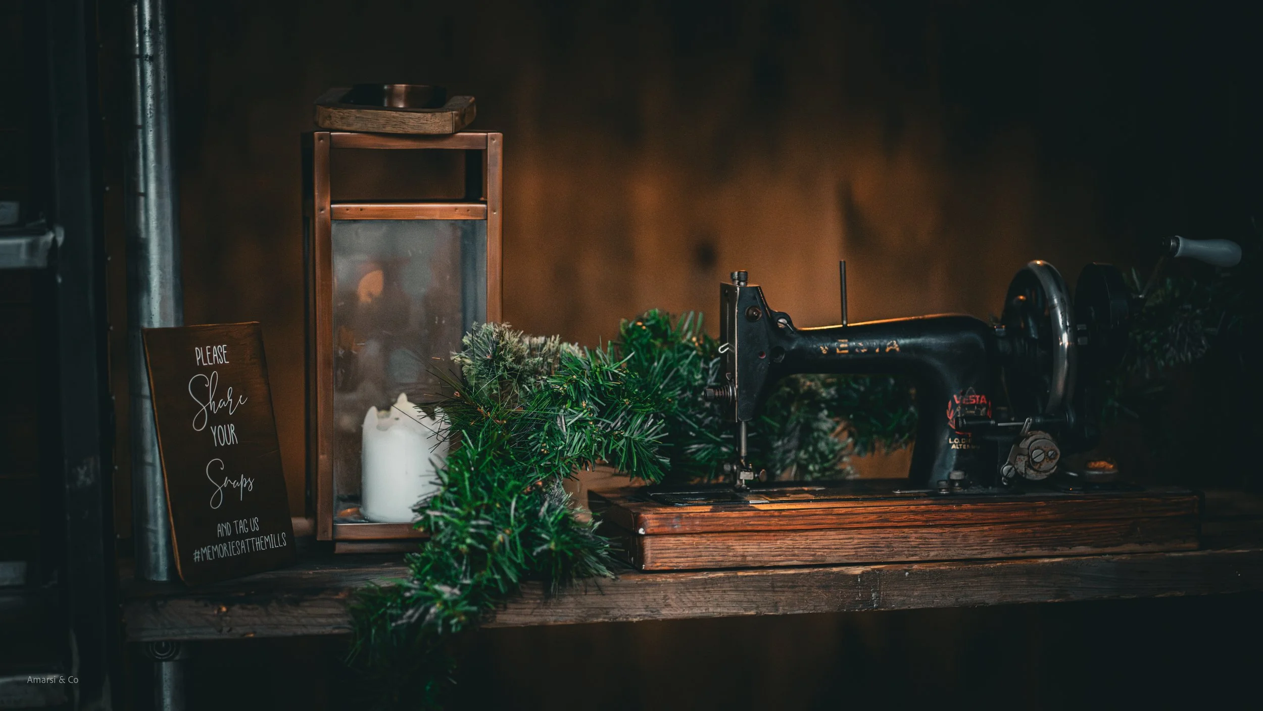 Vintage manual meat grinder on a wooden counter adorned with green holiday garland, with a candle in a glass container and a sign that reads, "Please share your scraps and tag us #memoriesatthemills" leaning against a lantern.