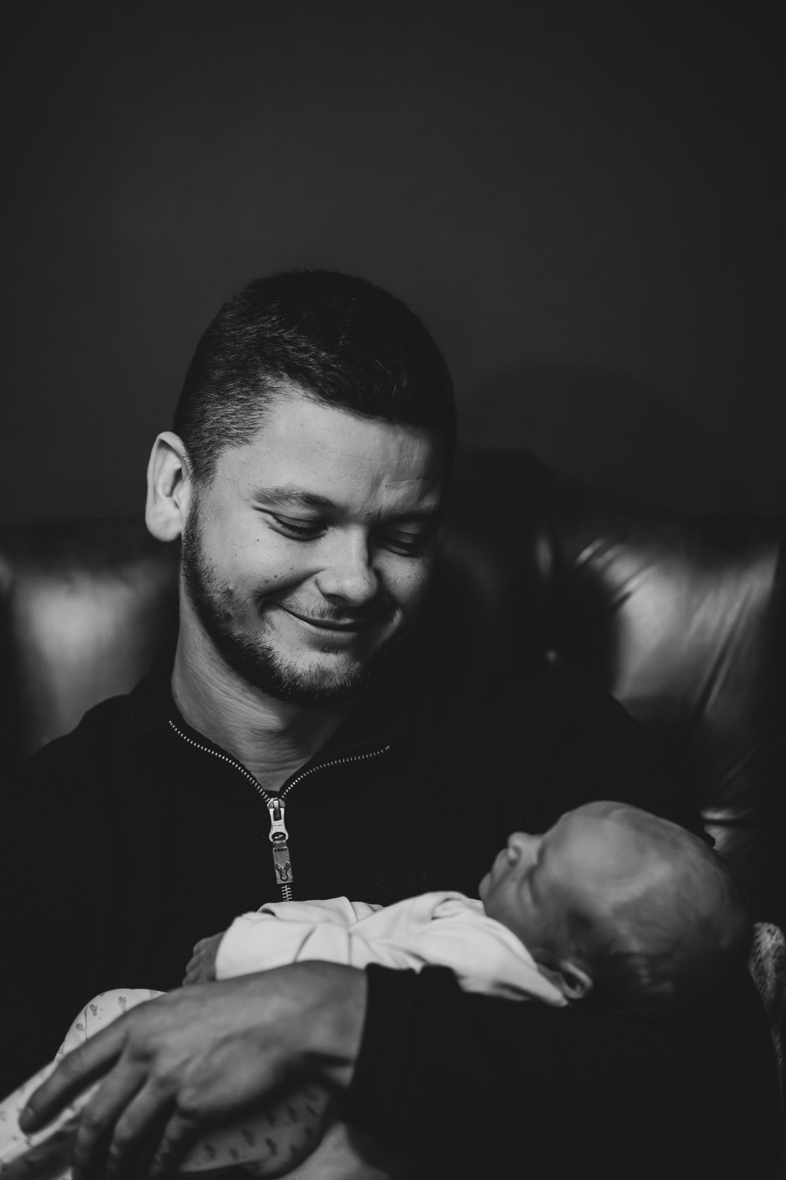 A man with short dark hair, a beard, and a smile on his face, is looking down at a baby he is holding gently. The background is dark, and only part of the man's arm and the baby's head are visible.
