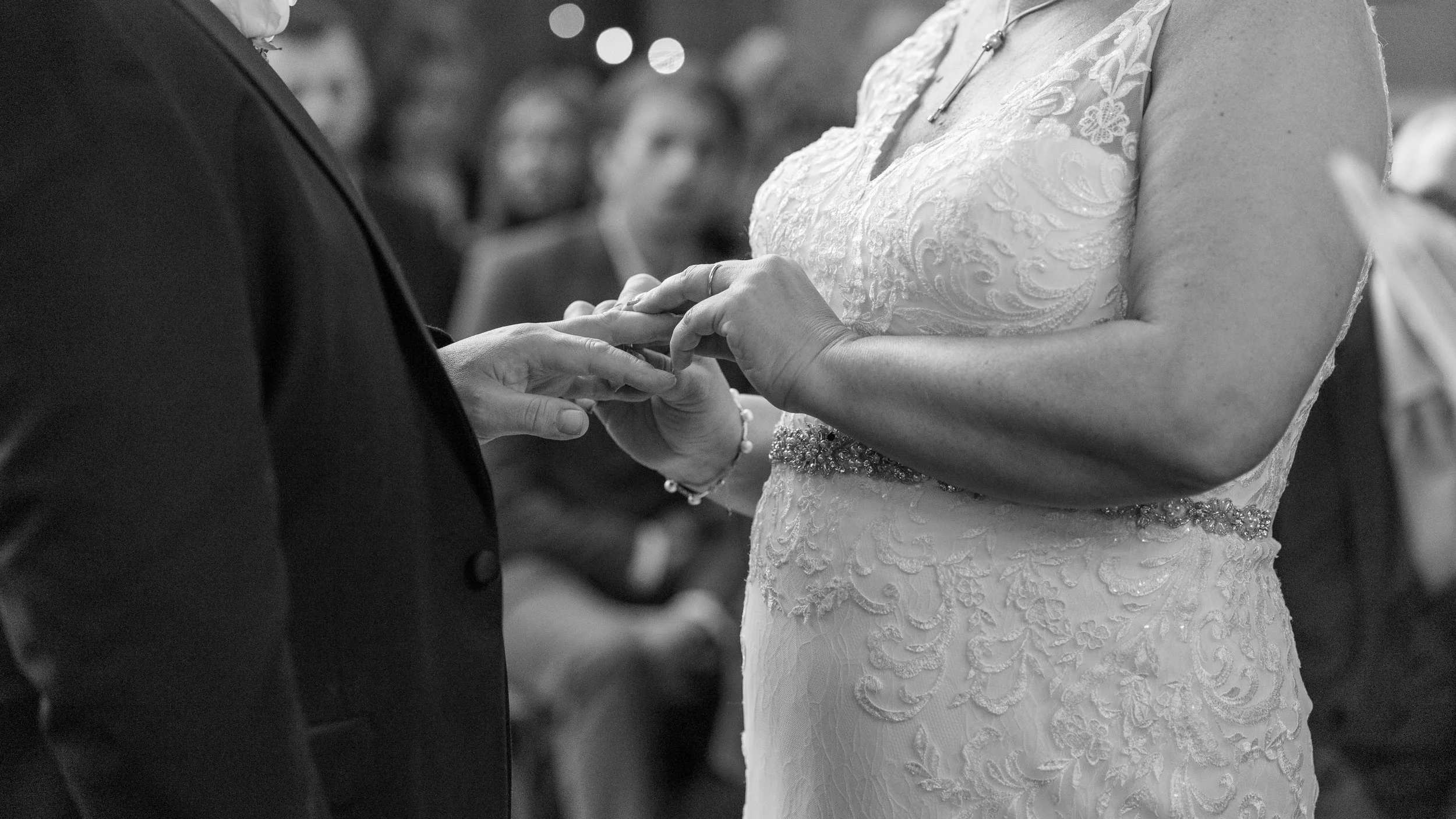 A bride and groom exchange rings during their wedding ceremony, with guests watching in the background.